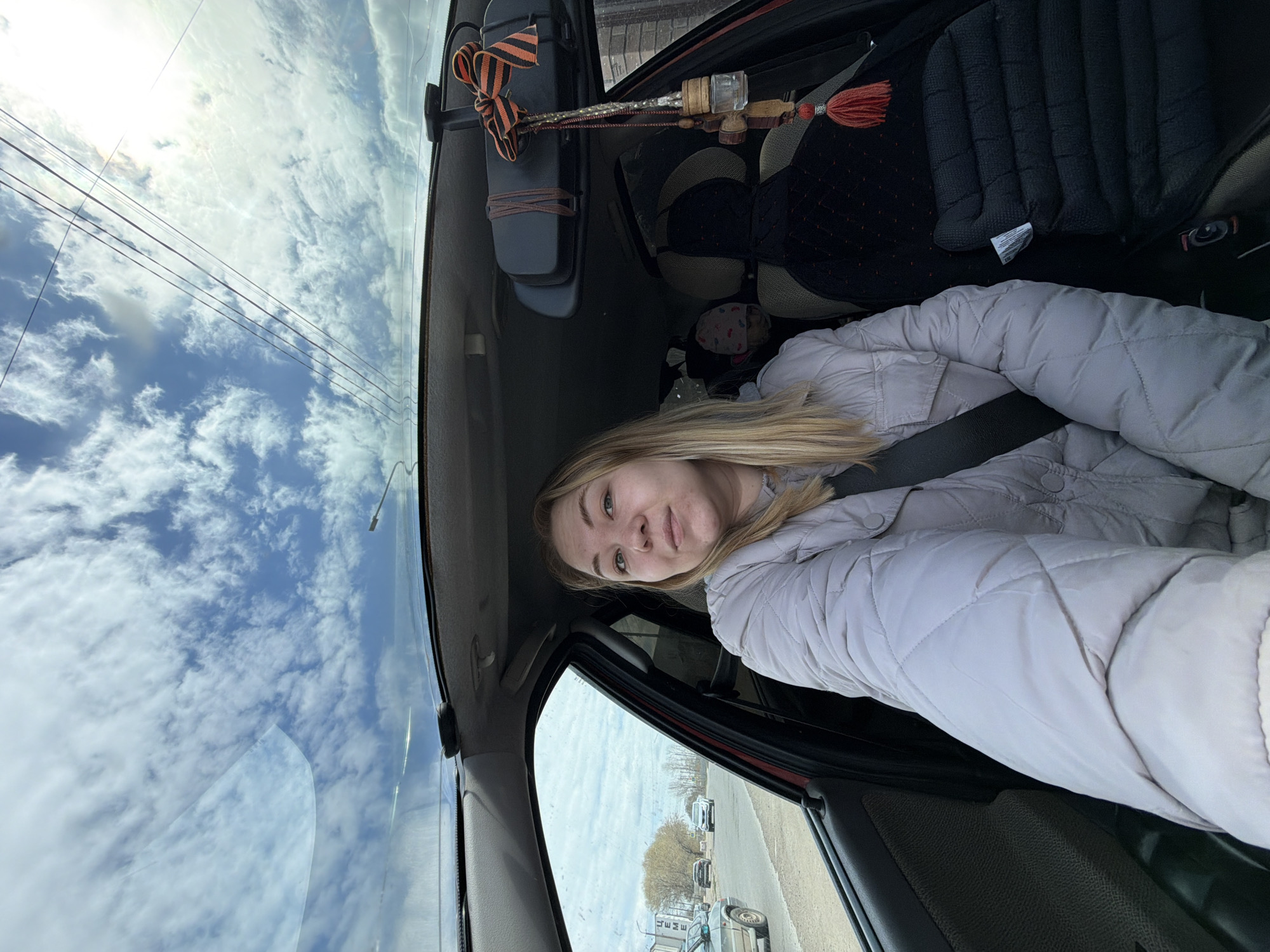 A woman with blonde hair is sitting in the driver's seat of a car, wearing a light-colored quilted jacket and a seatbelt. The view through the windshield shows a street with cars parked along the side and buildings in the background. The sky is visible with white clouds and blue patches. A St. George ribbon and a decorative wooden object with tassels hang from the rearview mirror. Based on the presence of the St. George ribbon, the image is most likely taken in Russia. There is no indication of the image being taken in Chaikovsky specifically.