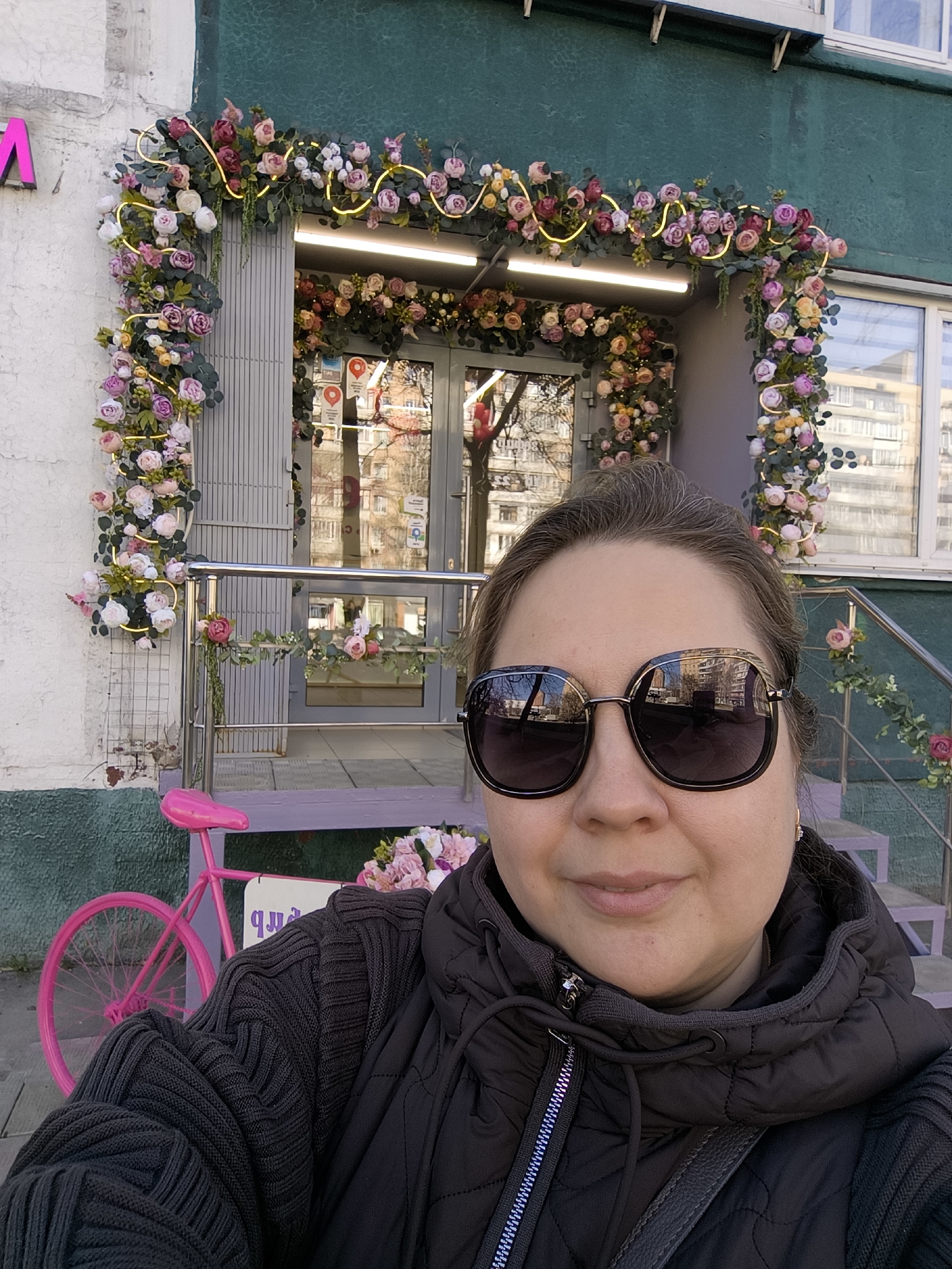 A medium shot depicts a woman in sunglasses and a black hooded jacket, posing in front of a flower-adorned storefront in Khimki, Russia. The entrance is decorated with an arch of flowers and fairy lights, creating a visually striking frame. A pink bicycle is partially visible on the left side of the frame. The building features green paint above the entrance and a white facade on the left side. The reflection in the sunglasses shows surrounding buildings, indicating a cityscape.