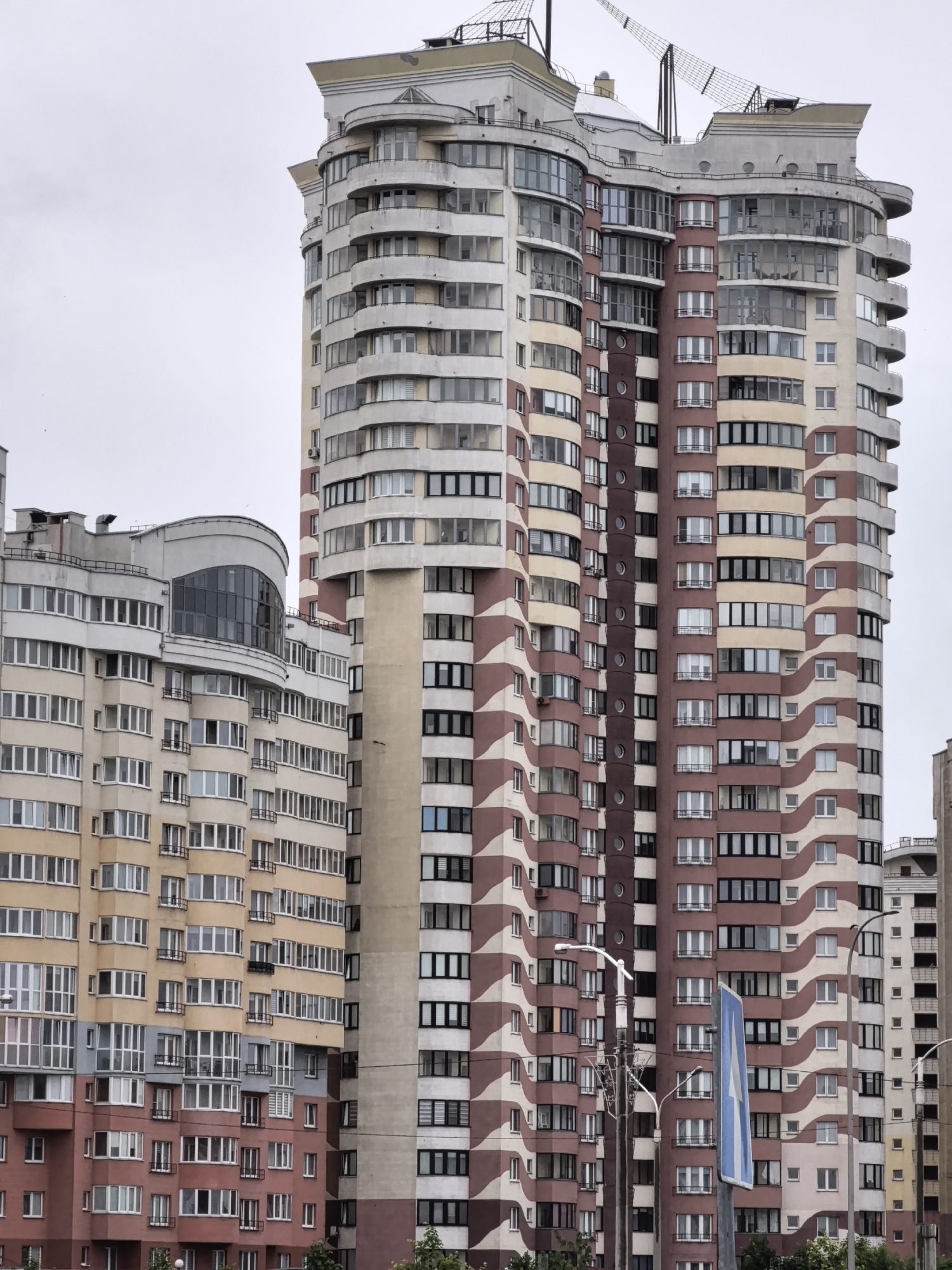 A medium shot captures a cluster of residential buildings against a pale, overcast sky. The central building is tall and curvilinear, featuring a pattern of alternating maroon and cream-colored wavy bands. It is adorned with numerous windows and balconies, some of which are curved to match the building's shape. To the left of this building, another residential structure is visible, with a facade of cream and pinkish-red lower levels, also featuring many windows. A third, narrower building, with a lighter cream color and darker horizontal bands, stands to the right of the central structure. Various streetlights and a blue directional sign are present in the foreground. The image depicts an urban residential environment.