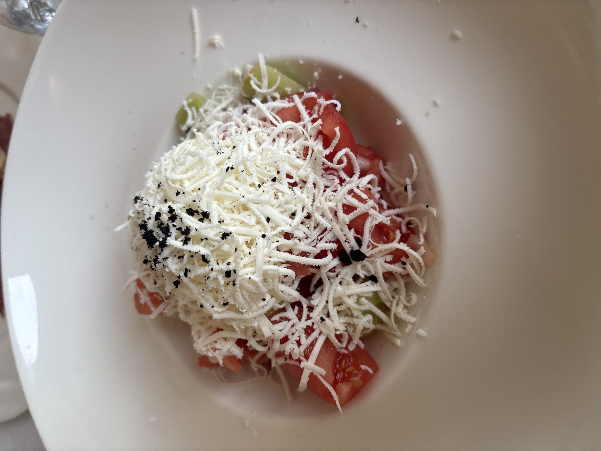 A close-up, overhead shot shows a serving of chopped tomatoes and cucumbers topped with grated white cheese and black specks, possibly pepper or herbs, presented in a wide, white ceramic bowl. The dish is likely located at a restaurant in Varna, Bulgaria, given the context of the provided information. No people are visible in the image. The background is blurred, suggesting an indoor setting, possibly a dining area.