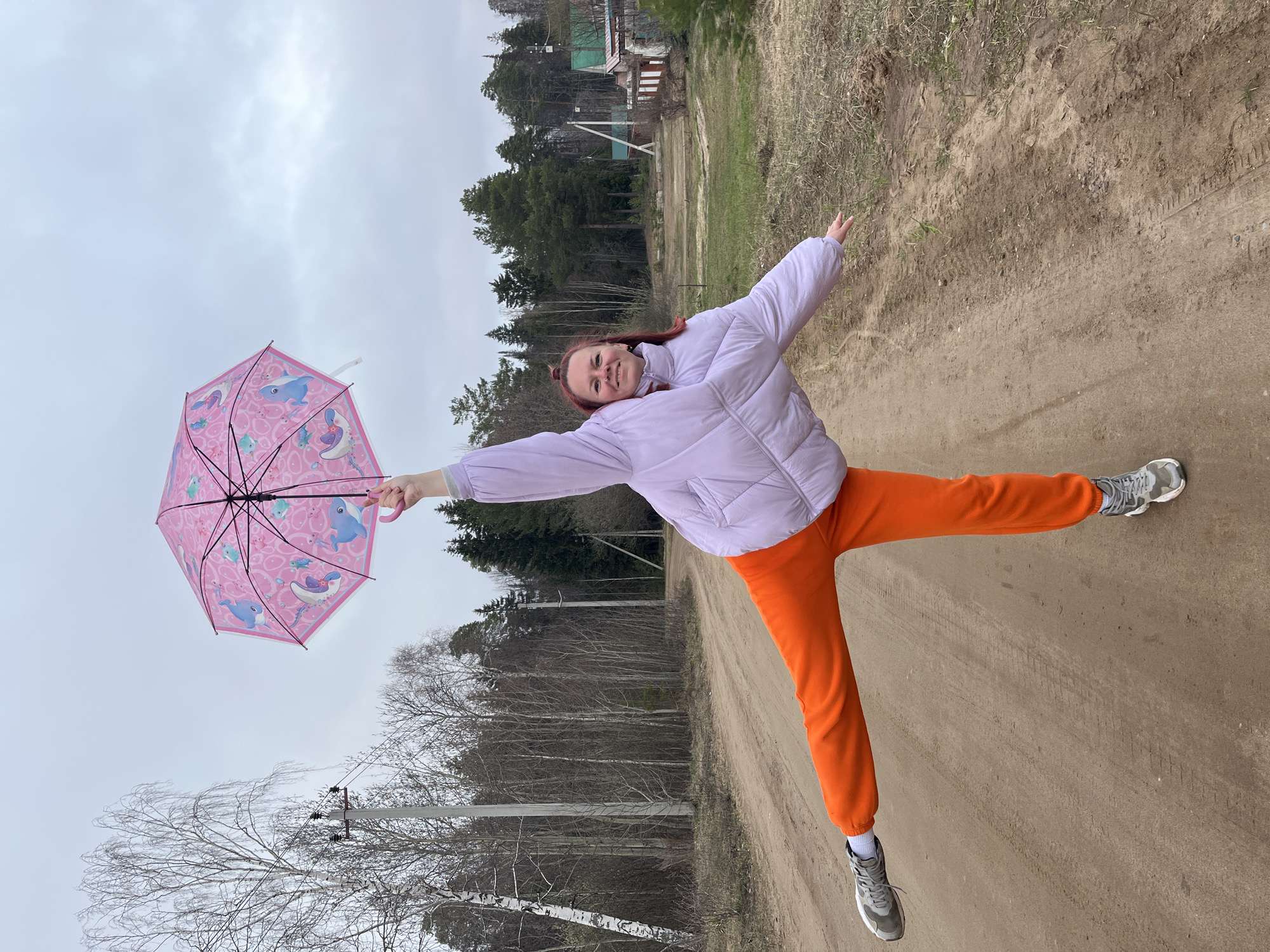A cheerful young woman is captured in mid-air, balancing playfully on one leg with her arms outstretched, on a dirt road in a rural setting near Chaikovsky, Russia. She has reddish-brown hair pulled back into a bun and is dressed in a light purple puffy jacket, bright orange sweatpants, and grey athletic shoes. In her left hand, she holds a vibrant pink umbrella adorned with cartoon marine animals, including dolphins, whales, and fish. Her face expresses pure joy with a wide, radiant smile.
The scene unfolds on an unpaved dirt road, marked by visible tire tracks. The background to her left features a sparse forest composed of leafless deciduous trees, such as birches, and some evergreen pines, alongside a utilitarian power pole with wires. To her right, the landscape consists of brush and distant trees, with glimpses of simple, rustic houses nestled among the foliage. The sky above is entirely overcast and cloudy, suggesting a cool, gray day without direct sunlight.