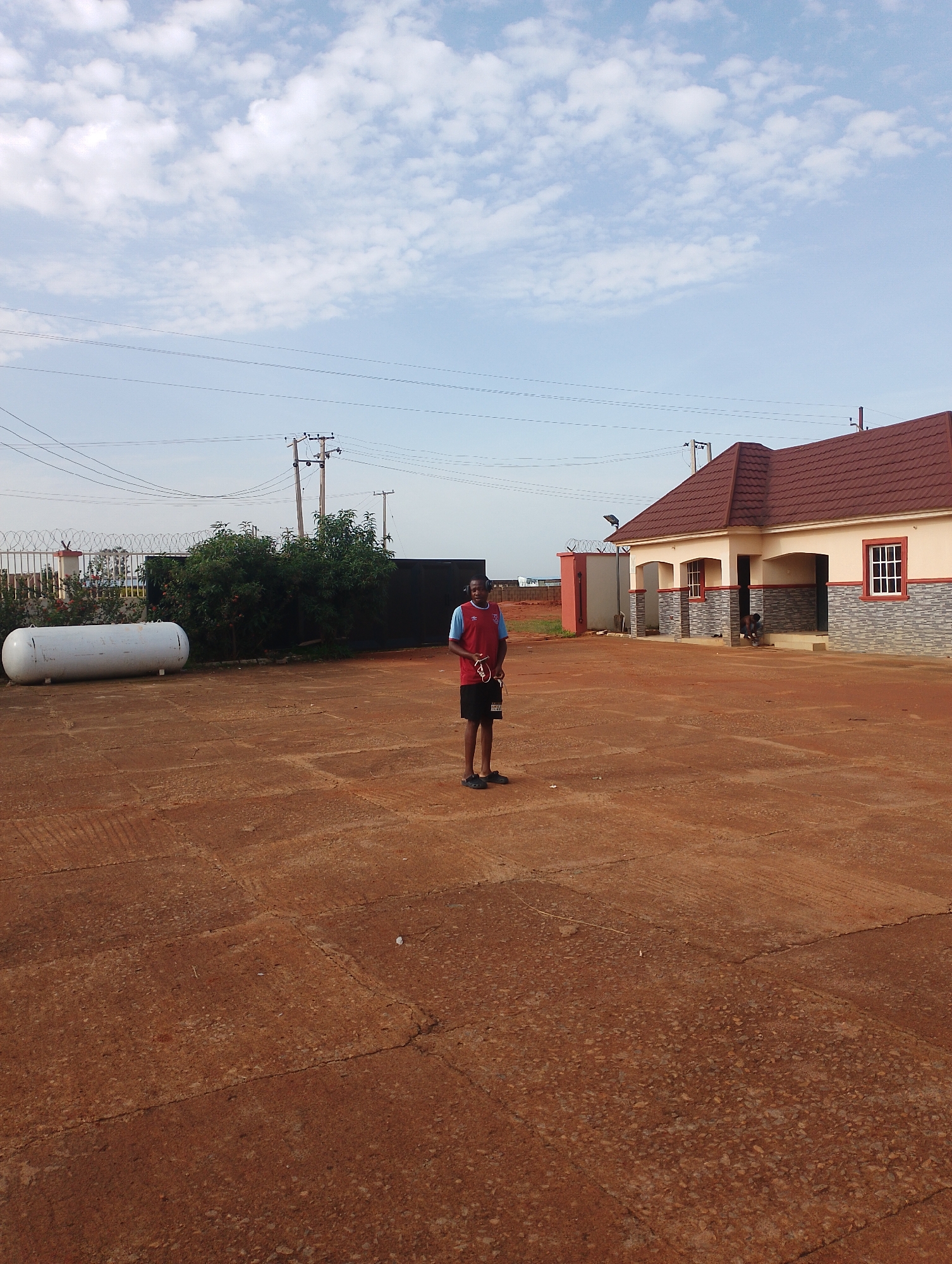 The image depicts an outdoor scene in Jos, Nigeria, under a partly cloudy sky. In the foreground, a young Black male stands in the center of a red, paved area that appears to be a courtyard or open ground. He is wearing a maroon and blue athletic shirt with a V-neck and black athletic shorts. He is holding a red and black object, possibly a sports item. To his left, a large white cylindrical tank is positioned on the ground. In the background, a building with a brown tiled roof and cream-colored walls is visible. The building features arched openings supported by pillars and has a dark-colored entrance. To the left of the building, there is a fence with barbed wire and some greenery, including trees and flowering bushes. Power lines and utility poles are also present in the background, stretching across the frame. The ground surface is predominantly red earth with some paved sections showing cracks and tire tracks. The overall impression is one of a semi-developed urban or semi-urban environment.