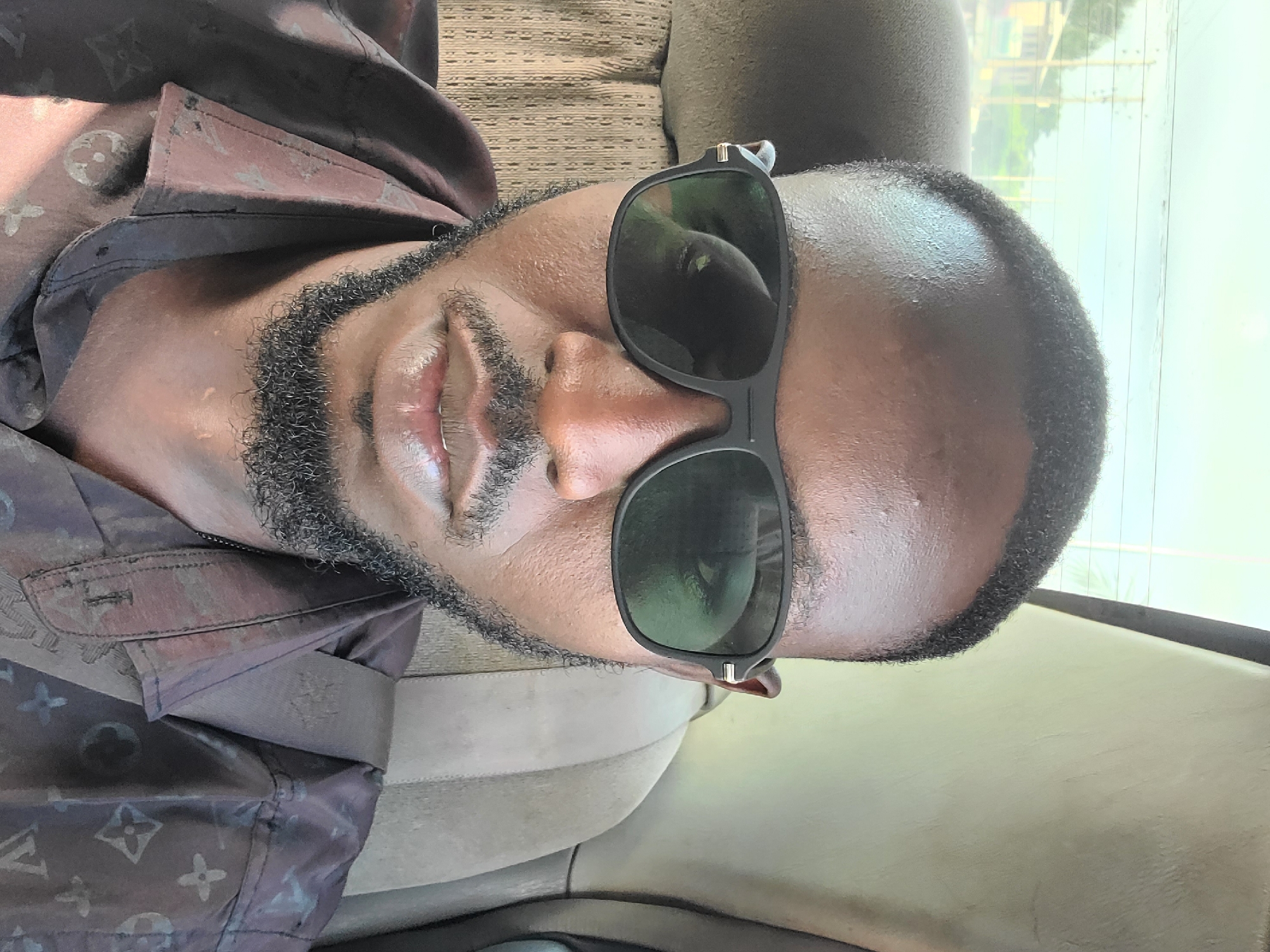 A close-up, eye-level selfie of a Black man wearing sunglasses and a patterned shirt, taken inside a vehicle. The man has short, dark hair, a beard, and is looking directly at the camera. The background consists of the interior of the car, including a headrest and a window showing a blurred outdoor scene with buildings and utility poles. The lighting suggests it is daytime. The overall impression is a casual, personal photograph.
