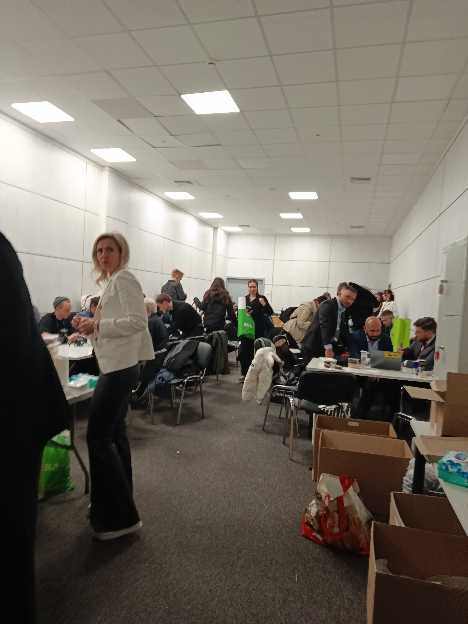 The image appears to be taken indoors, potentially in a large hall or office space, possibly in Khimki, Russia. A group of people is present, seated around tables. Some individuals are working on laptops, while others are engaged in conversation or eating. A woman in a white blazer and dark pants is looking towards the camera. There are several cardboard boxes in the foreground, suggesting the presence of supplies or items being packed/unpacked. The environment seems to be well-lit with fluorescent ceiling lights. The scene suggests a collaborative workspace or a gathering for a specific purpose, possibly related to aid or assistance, based on the presence of boxes.