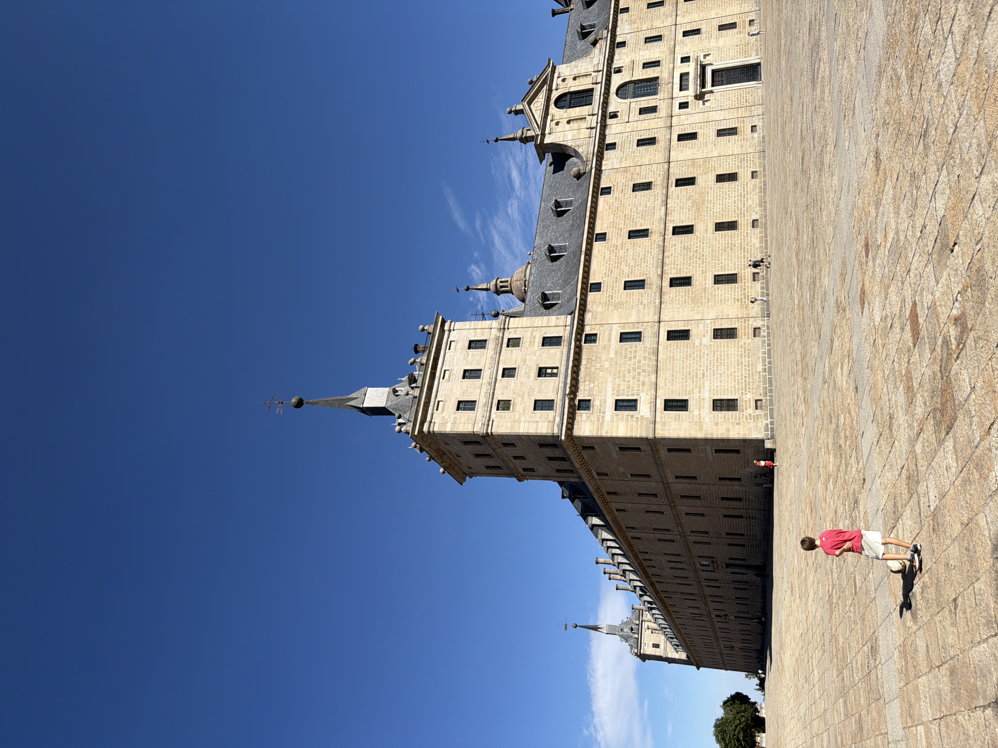 A wide-angle, eye-level, outdoor shot shows the monumental facade of the Royal Site of San Lorenzo de El Escorial under a clear, bright blue sky. The massive sandstone building, characterized by its imposing architecture and symmetrical design, dominates the frame. Its long, extended wings are adorned with numerous dark rectangular windows and several prominent towers topped with spires and cross finials. The building features a grand entrance with a dark, imposing doorway. In the foreground, a young boy, wearing a red t-shirt and white shorts, stands on a vast, paved courtyard composed of large, square stone tiles. He is facing away from the camera and appears to be looking at a soccer ball at his feet, casting a distinct shadow on the ground. The courtyard is expansive and mostly empty, with a few other distant figures and trees visible along the edge of the building and in the far background. The overall scene conveys a sense of grandeur and historical significance, contrasted with the solitary presence of the child.