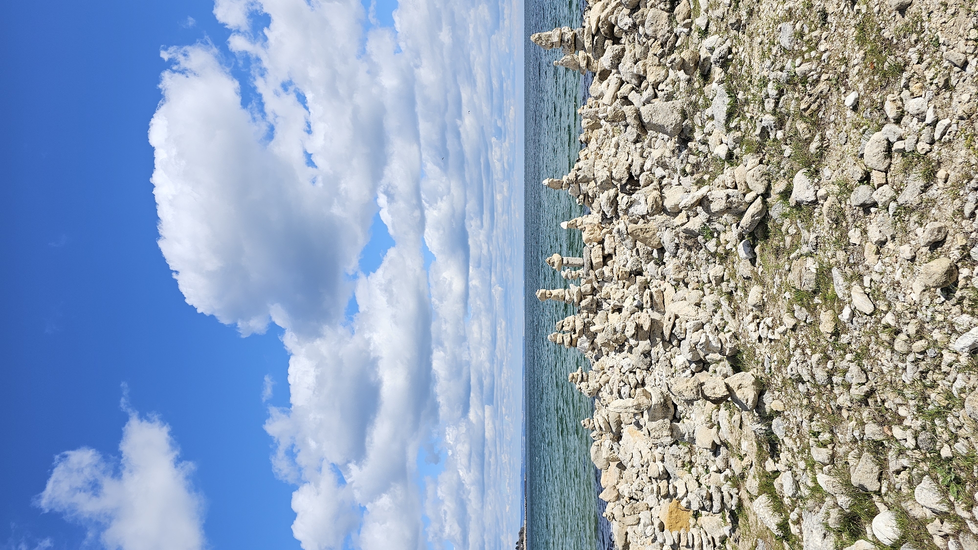 The image shows a rocky beach with stacked stones on the shoreline, adjacent to a body of water. The sky is visible above the water, featuring both blue sky and clouds. The land portion is populated by stones of various sizes and some sparse vegetation. This scene is located in Varna, Bulgaria.
