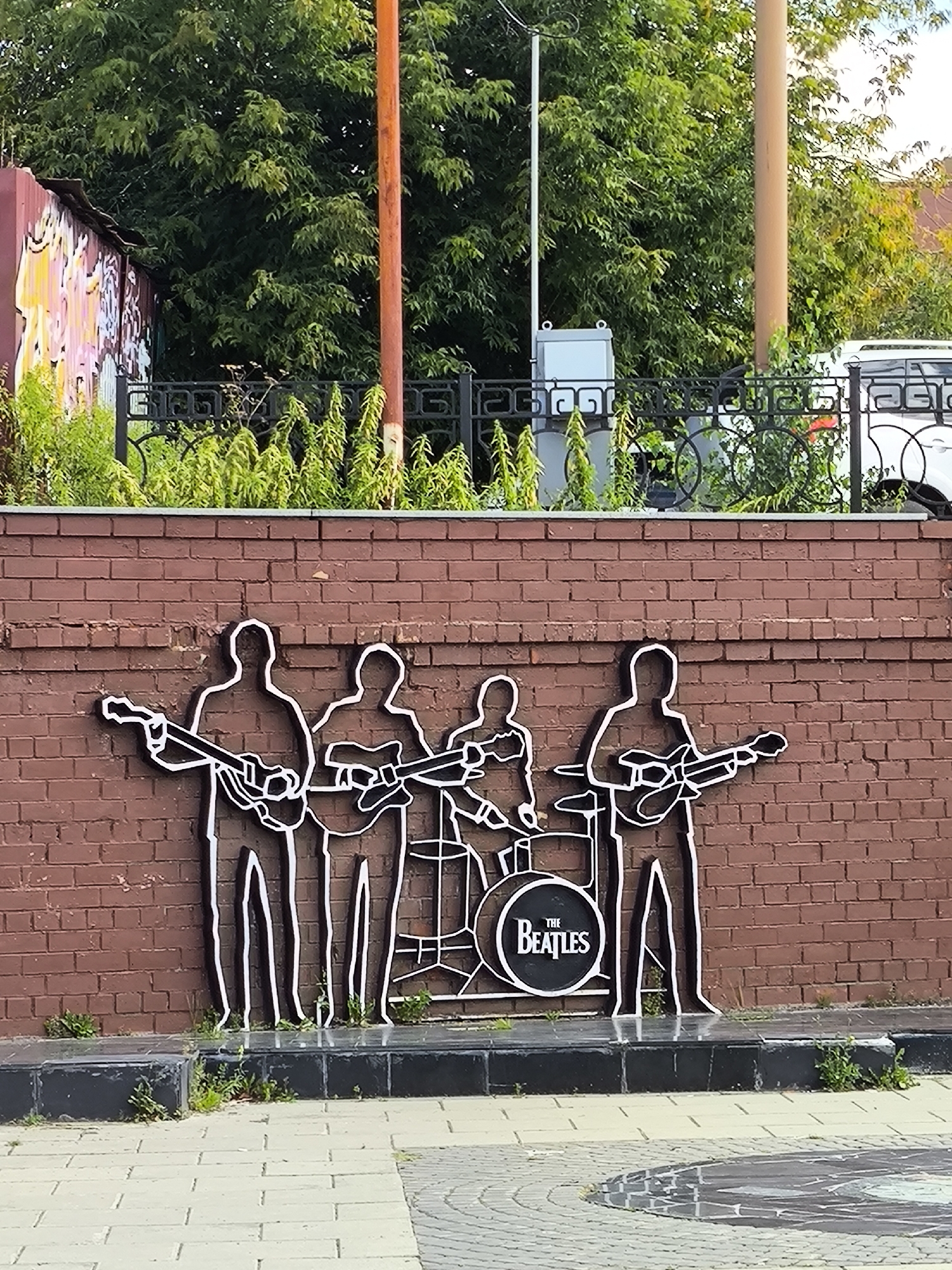 The image shows a white outline art installation of the band The Beatles performing on a brick wall. The figures are depicted playing guitars and drums. The band's logo, "The Beatles," is visible on the bass drum. The installation is located in Yekaterinburg, Russia, at the intersection of Malysheva Street and the embankment of the Iset River. The background includes trees, a fence, and a utility box.