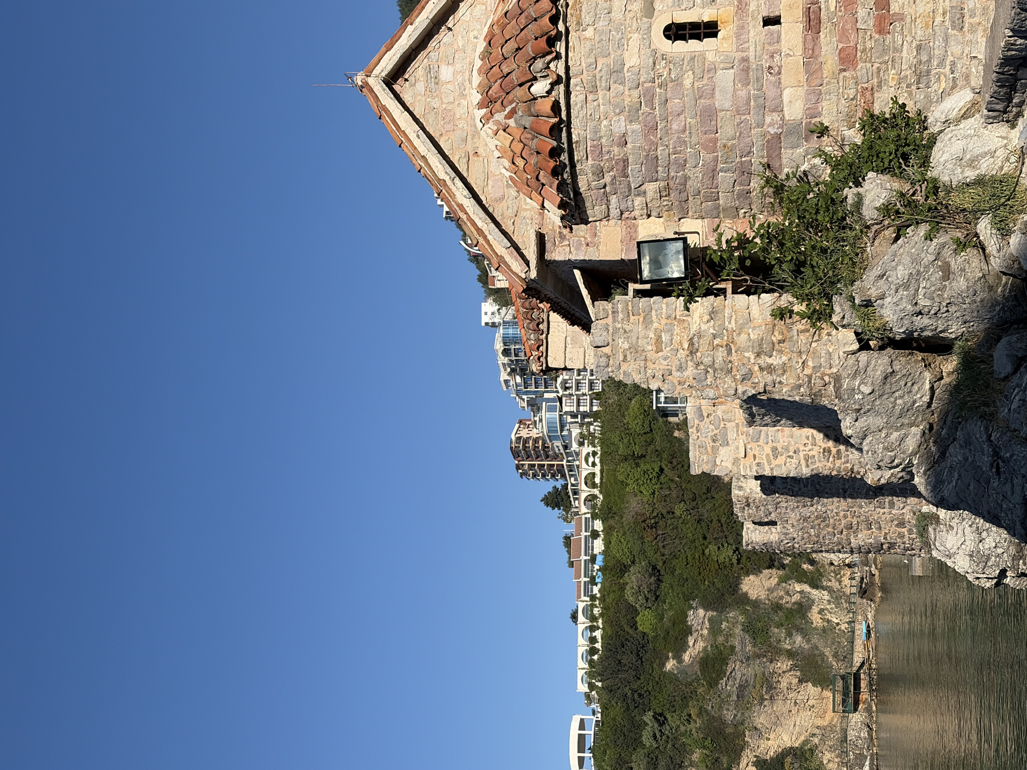 The image captures a section of the historic Sveti Stefan island connected to the city of Budva, Montenegro. The foreground features a stone structure with a tiled roof and arched window openings, typical of Mediterranean architecture. Adjacent to the structure, there are rocky formations with vegetation. In the background, modern buildings are visible. A body of water is also visible. The sky is clear and blue.