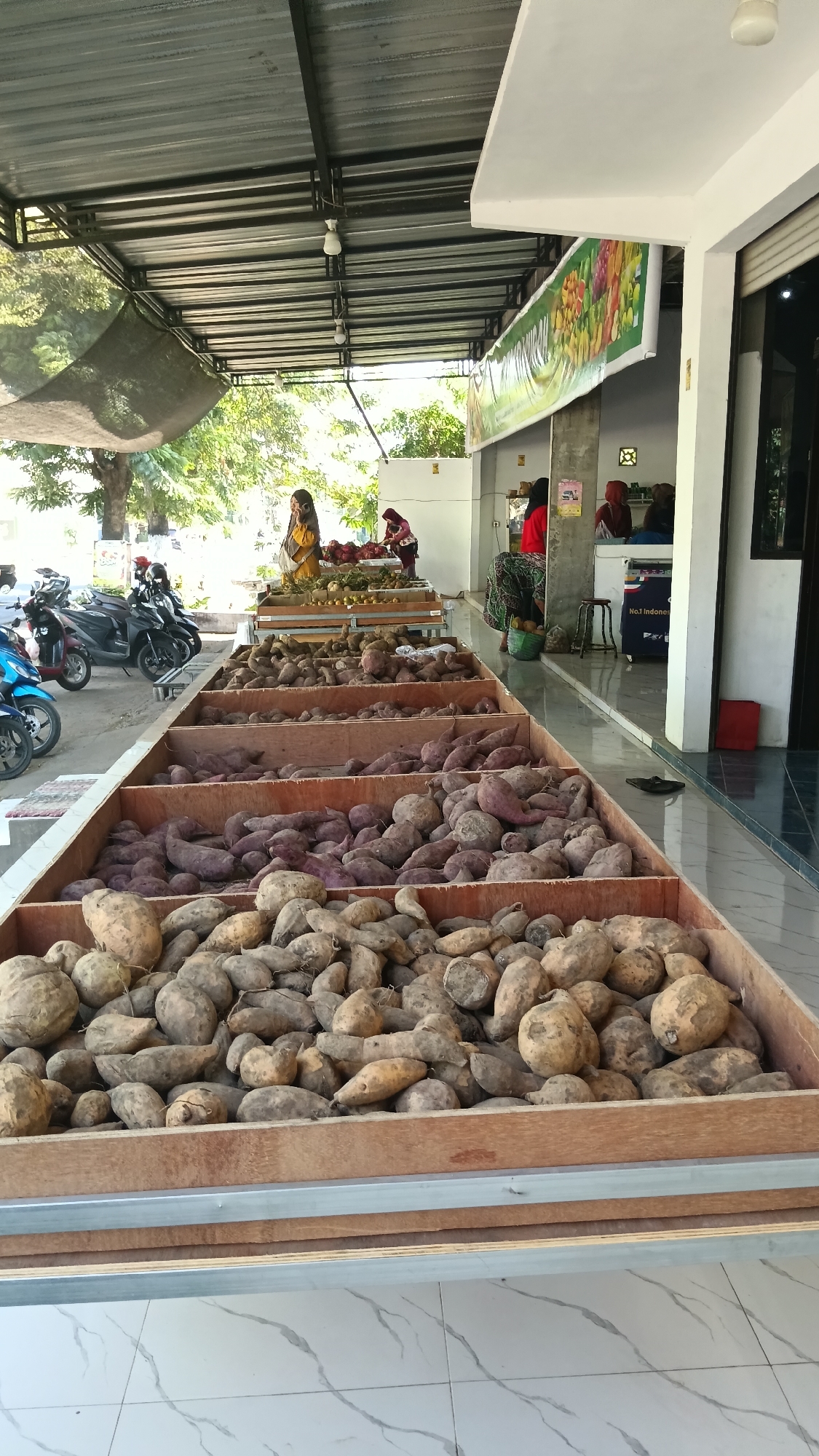 The image depicts an outdoor market scene in Pule, Indonesia, featuring a large display of root vegetables, likely sweet potatoes, arranged in wooden crates. In the foreground, several long, shallow wooden crates are filled with these root vegetables, varying in color from light tan to deep purple. Behind these, a raised display table holds more produce, including oranges and possibly other root vegetables.
Several people are present in the background. To the left, three motorcycles are parked. Further back, under a covered structure supported by metal beams, a woman in a yellow headscarf is standing near the produce display, potentially interacting with customers. Another woman in a red headscarf is also visible at the produce stand. To the right, under the building's overhang, other individuals are seen, including a woman in a red top and patterned skirt sitting on a stool, and others who appear to be vendors or customers. The environment suggests a typical Indonesian market setting, with the open-air setup and the presence of motorcycles for transportation.