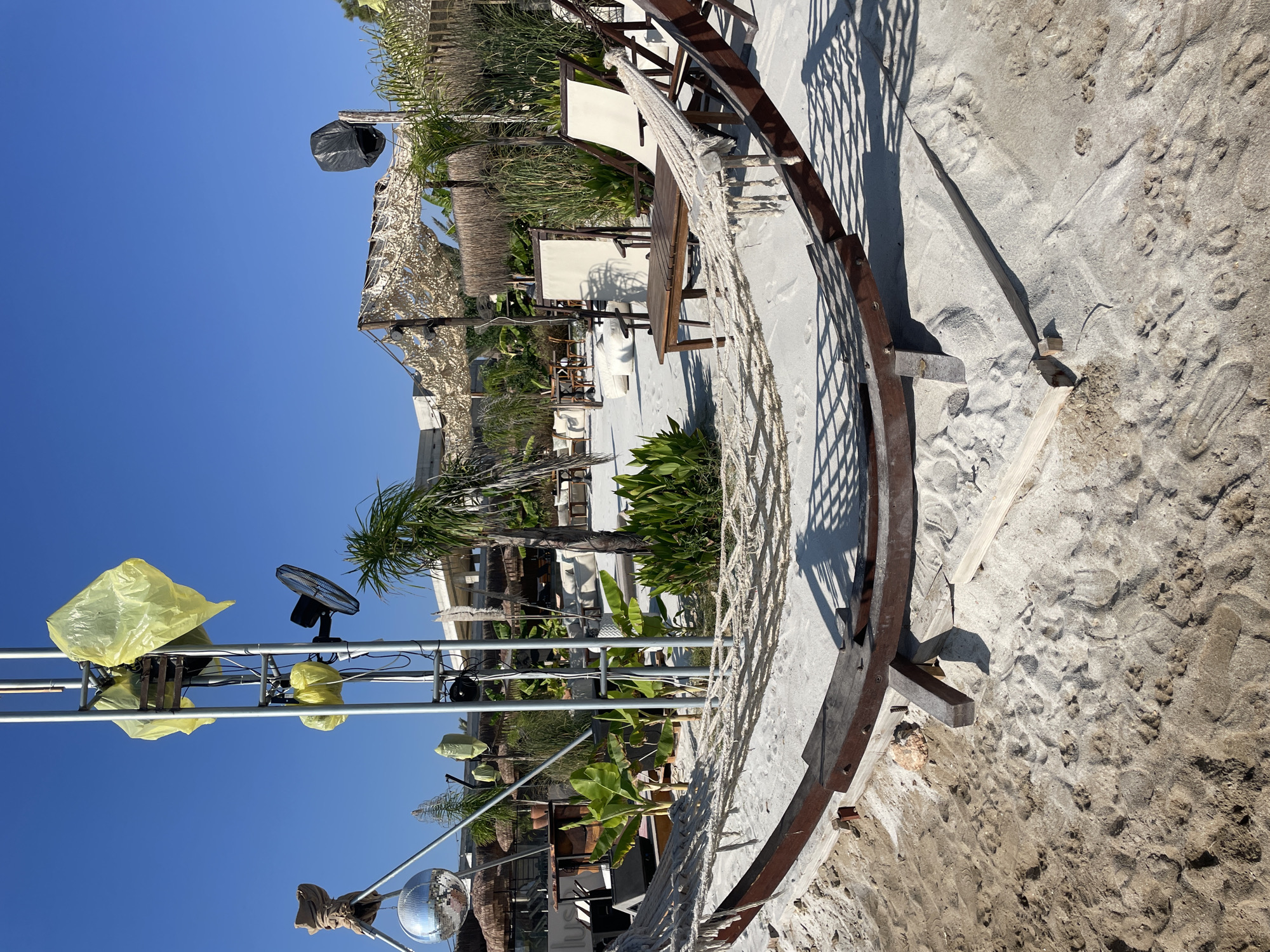 This image captures a sunny day at a beachside establishment in Trikomo, Northern Cyprus. In the foreground, a wooden hammock frame rests on the white sand, its ropes partially visible. Behind the hammock, a light-colored patio area is set up with white lounge chairs, a wooden table, and various potted plants, including banana trees and tall grasses. A thatched-roof structure with decorative netting provides shade. To the left, a metal pole supports a disco ball, a fan, and several yellow plastic bags covering equipment, suggesting preparation or protection from the elements. Further back, the beach extends to a building with a modern architectural design, partially under construction. The sky is a clear, bright blue, indicating good weather.