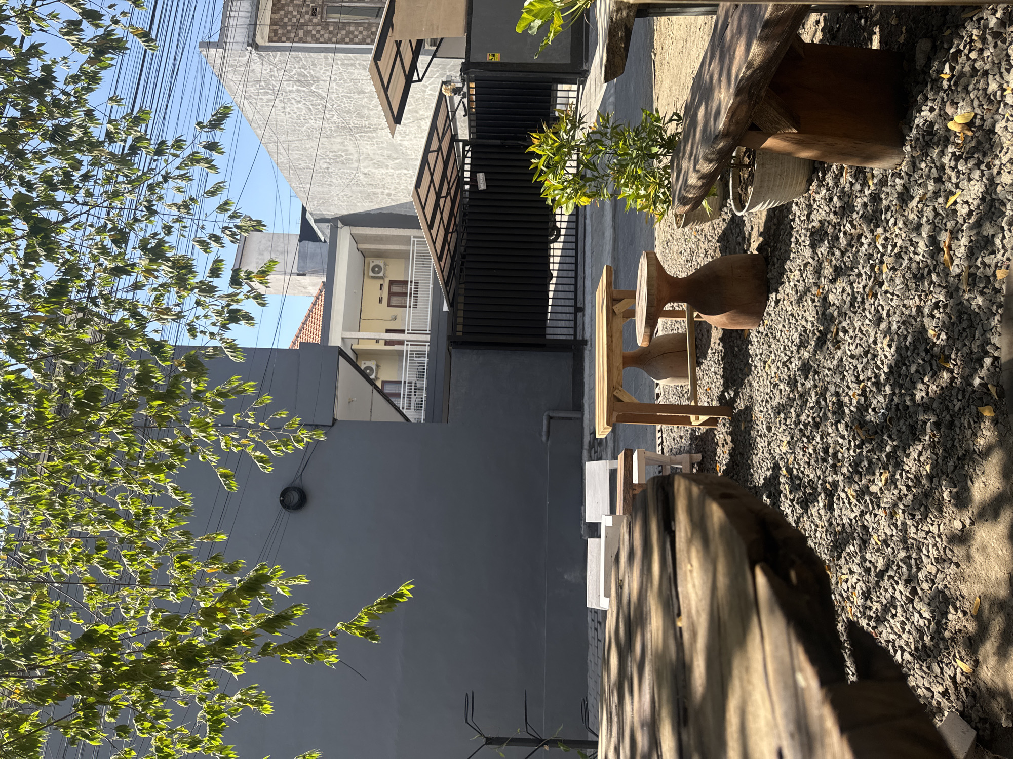 The image captures an outdoor seating area of a cafe in Denpasar, Indonesia. The foreground is dominated by a rustic wooden table with a weathered surface, partially obscuring the view of the rest of the scene. The ground is covered in small gray gravel, with scattered dry leaves and shadows cast by overhead foliage.
In the mid-ground, several pieces of outdoor furniture are arranged. There are two white chairs, a wooden table with a simple design, and a carved wooden stool. Further back, a black metal gate with a corrugated metal roof structure stands behind a small potted plant with green leaves. Beyond the gate, a building with a light-colored textured facade is visible, with a smaller structure in front of it featuring a white railing and windows. Another building with a dark gray wall is to the left, with a circular black object mounted on it. Overhead, power lines crisscross against a clear blue sky, and the branches of a leafy tree with green foliage extend into the frame from the top left, casting dappled shadows. The overall impression is a relaxed, open-air environment with natural light and a touch of rustic charm.