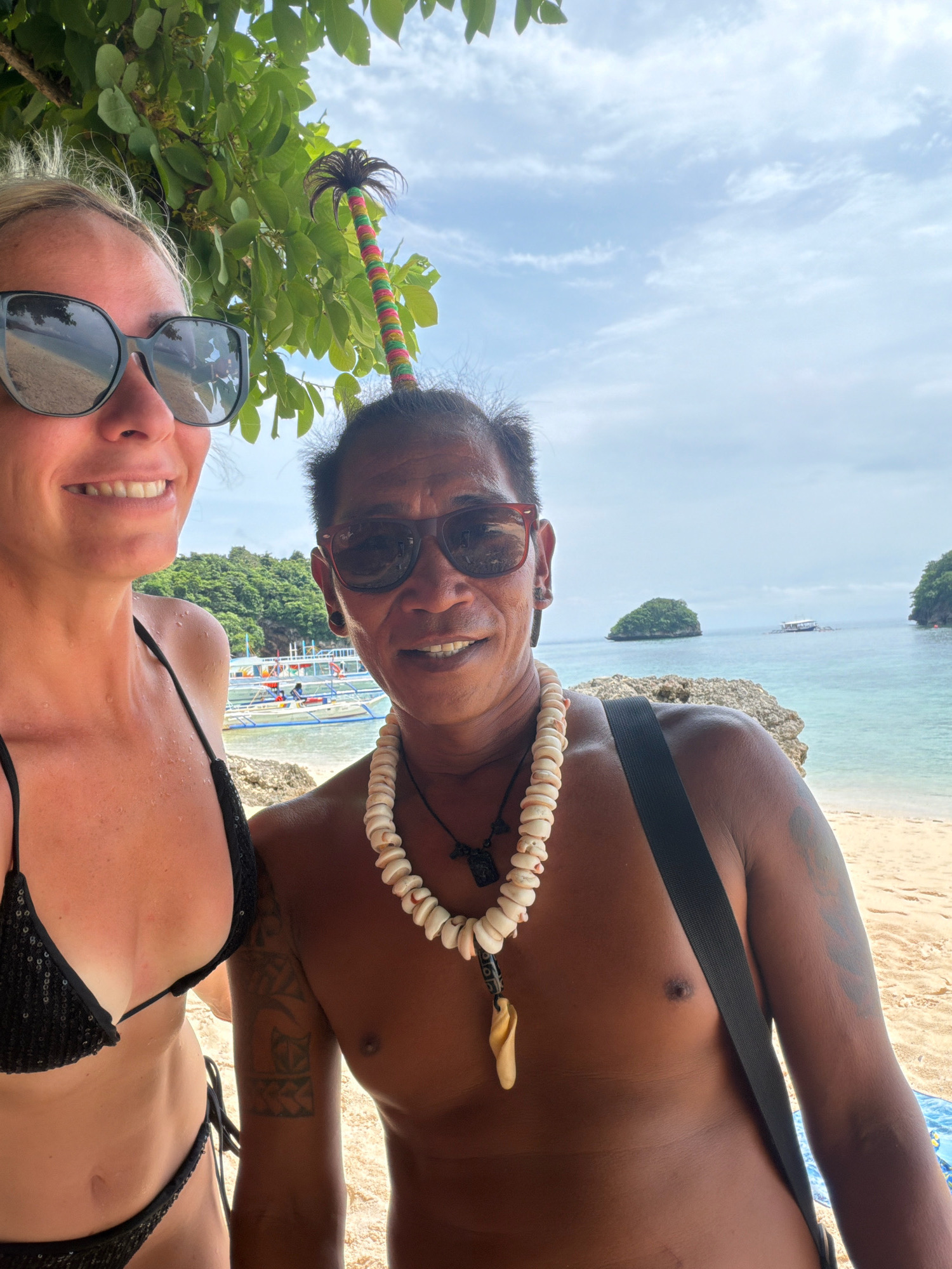Two individuals, a woman and a man, are posing for a selfie on a beach in Malay, Philippines. The woman, on the left, is wearing a black sequined bikini and large sunglasses. She has blonde hair and is smiling. The man, on the right, has dark hair, wears sunglasses, and a necklace made of shells. He is bare-chested, displaying tattoos on his arm. Behind them, the turquoise water of the ocean meets a sandy beach, with lush green islands in the distance. Several boats are visible near the shore, suggesting a tourist area. The sky is partly cloudy.