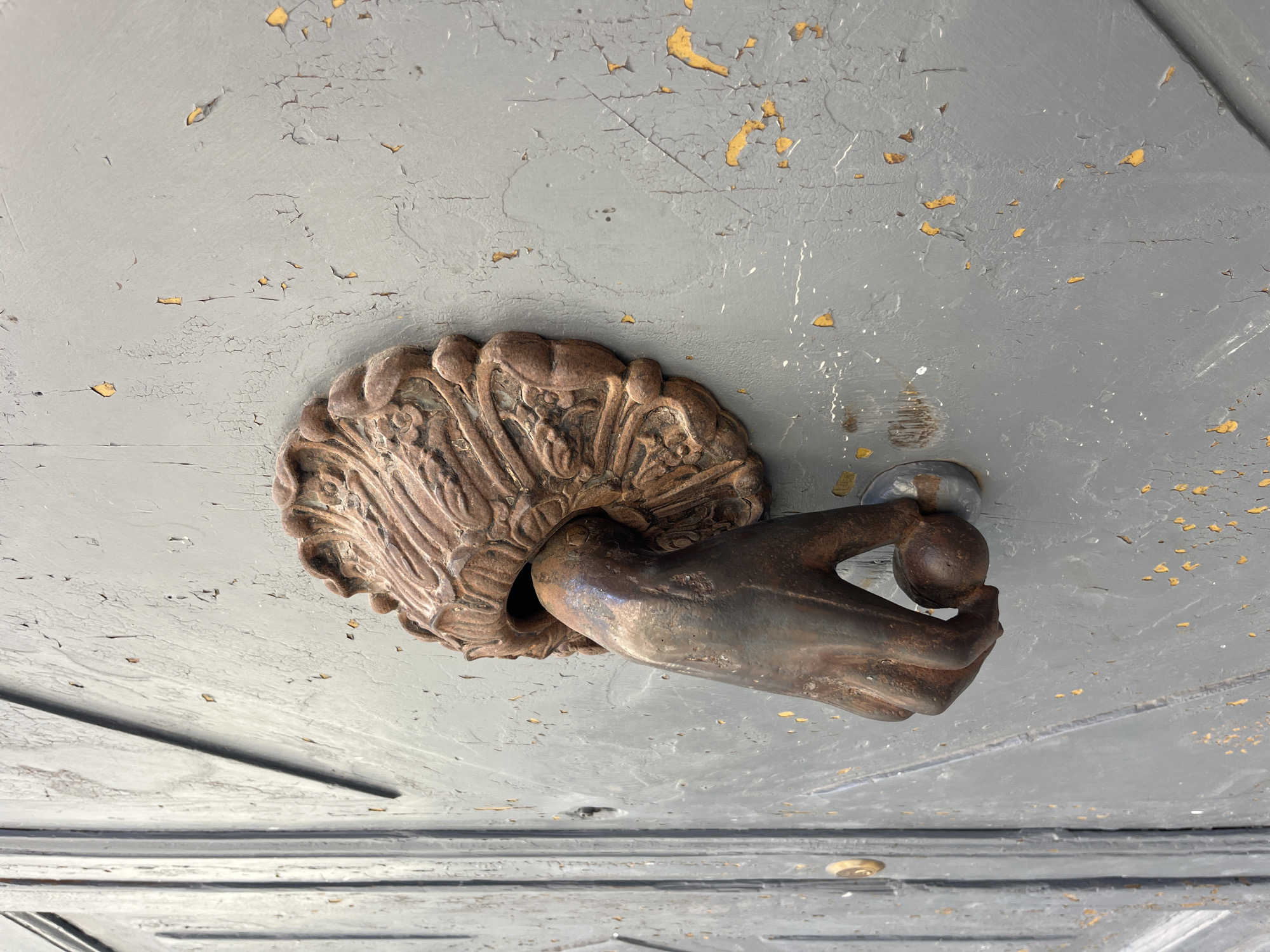 The image shows a close-up of a weathered, cast-iron door knocker shaped like a hand holding a ball. The knocker is mounted on a gray wooden door with peeling paint, revealing yellow undertones. The door has vertical panels and a visible keyhole on the left side. The knocker itself is ornate, with a decorative element at the top from which the hand emerges. The hand is detailed, showing fingers and the palm, and grips a spherical element. The scene appears to be an exterior shot, likely in Viterbo, Italy, given the architectural style and material of the door and knocker, suggesting an older building.