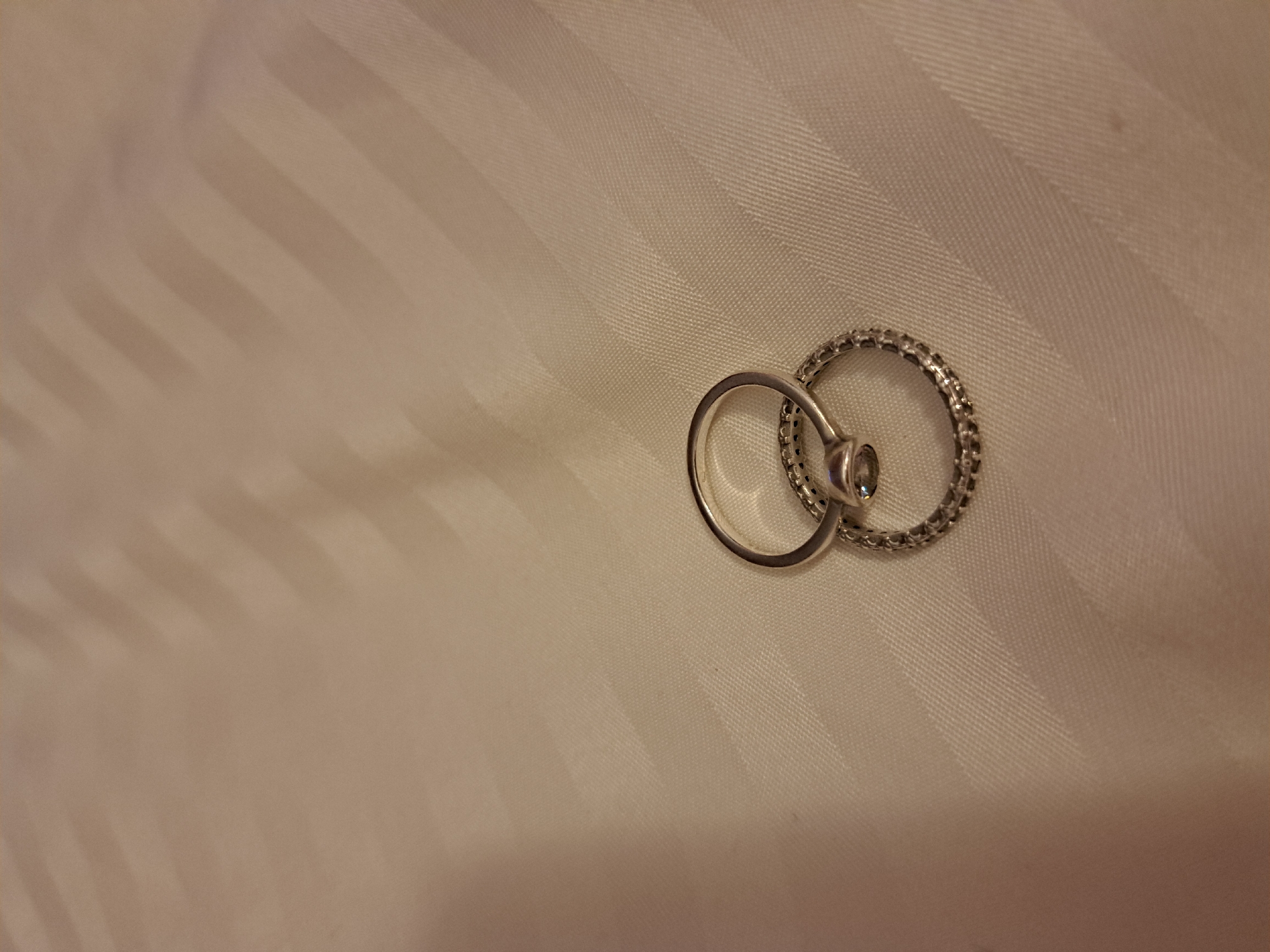 The image shows two intertwined rings on a striped surface. One ring is plain and silver, with a single small stone. The other ring is also silver and appears to be studded with small clear stones around its circumference. The surface is a light-colored fabric with a subtle striped pattern. There is no information to infer the image was taken in Negovan, Bulgaria, or any details about people, subjects, objects, actions, interactions, and the environment.
