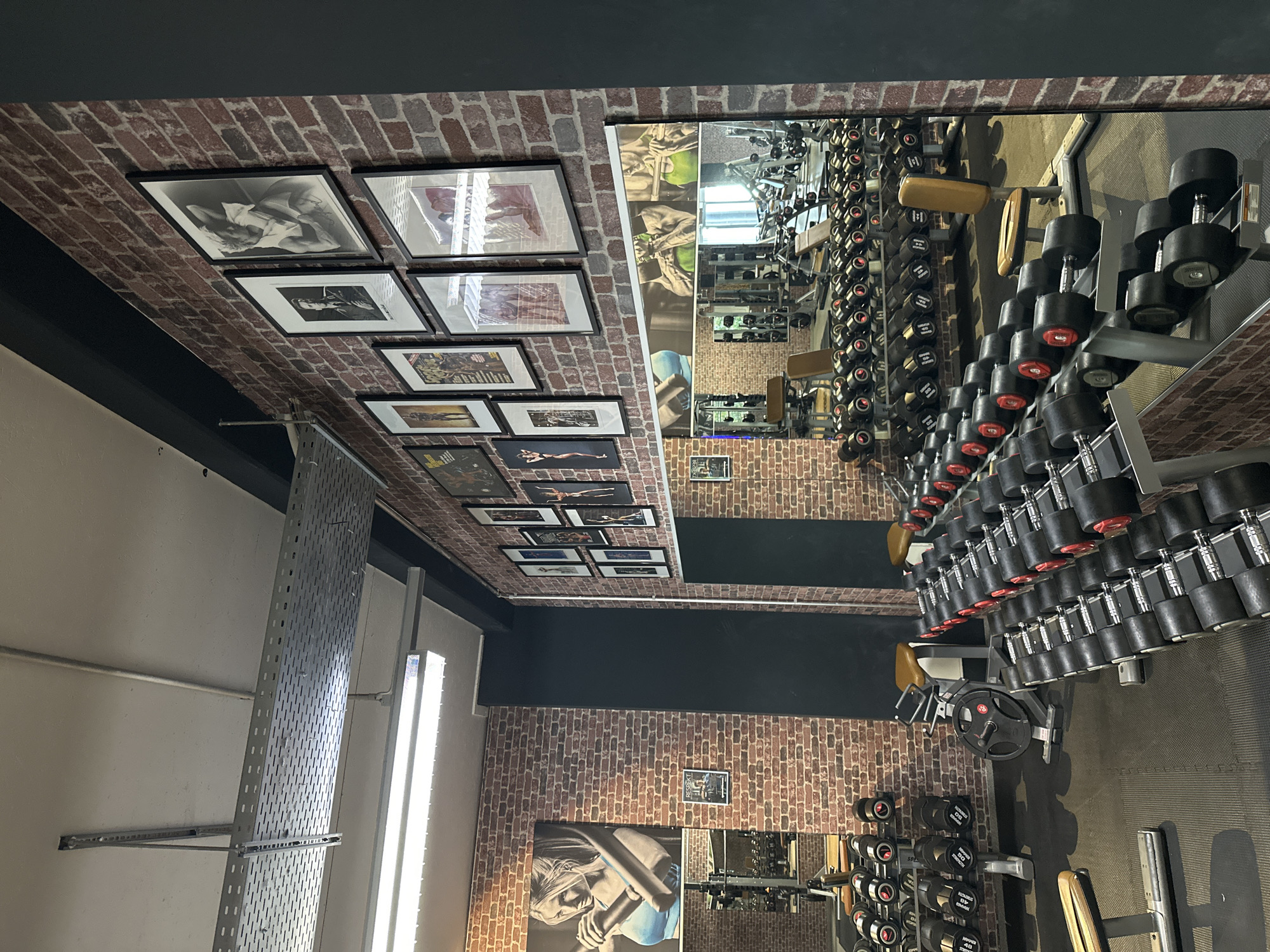 This is an interior shot of a gym, likely located in Brunnthal, Germany (due to the image title). The walls are clad in brick veneer and adorned with framed photographs. A large mirror reflects the gym's interior, showing rows of dumbbells organized on racks. Various weightlifting machines and equipment are present. There are no people visible in the immediate frame. The lighting appears to be a mix of artificial and natural sources. A metal shelving unit is visible on the left wall. The overall impression is of a well-equipped, modern fitness facility.