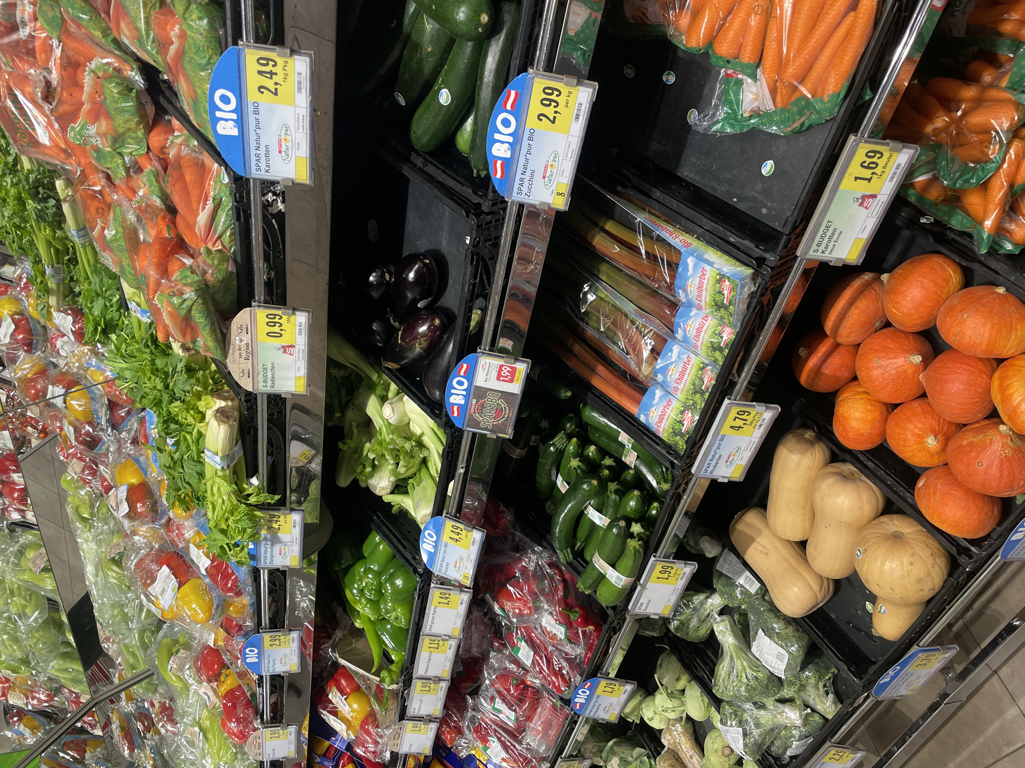 This is a photograph of a produce section in a grocery store, likely in Unknown, Unknown. The image shows various vegetables arranged in black display bins. From left to right, one can see an assortment of bagged bell peppers (red, yellow, green), celery stalks, bagged carrots, radishes, eggplants, fennel, zucchini, rhubarb, green bell peppers, more bagged bell peppers, and broccoli. Toward the right are butternut squash and small pumpkins. Each vegetable section has a price tag with information such as the brand ("S-BUDGET," "SPAR Natur pur BIO"), the name of the vegetable, and the price. Several price tags indicate "BIO," suggesting organic produce. The environment is well-lit and appears clean, typical of a grocery store setting.
