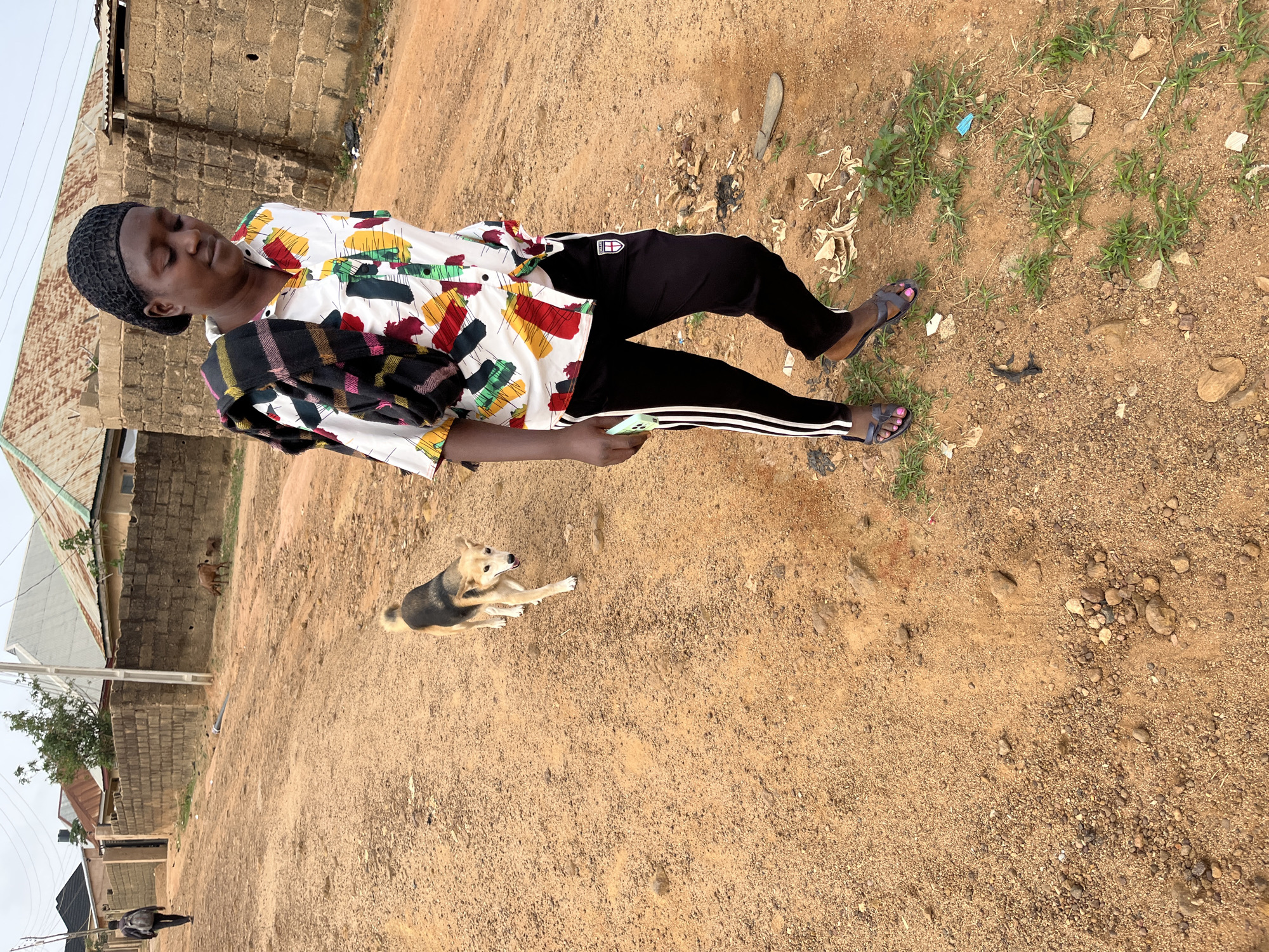 A person wearing a patterned shirt, black pants with white stripes, and sandals is standing on a dirt ground in an outdoor setting. The person is holding a smartphone. A dog is lying on the ground near the person. In the background, there are buildings, including a structure with a corrugated metal roof and brick walls. Additional dogs can be seen in the background near the buildings. The location is Jos, Nigeria.