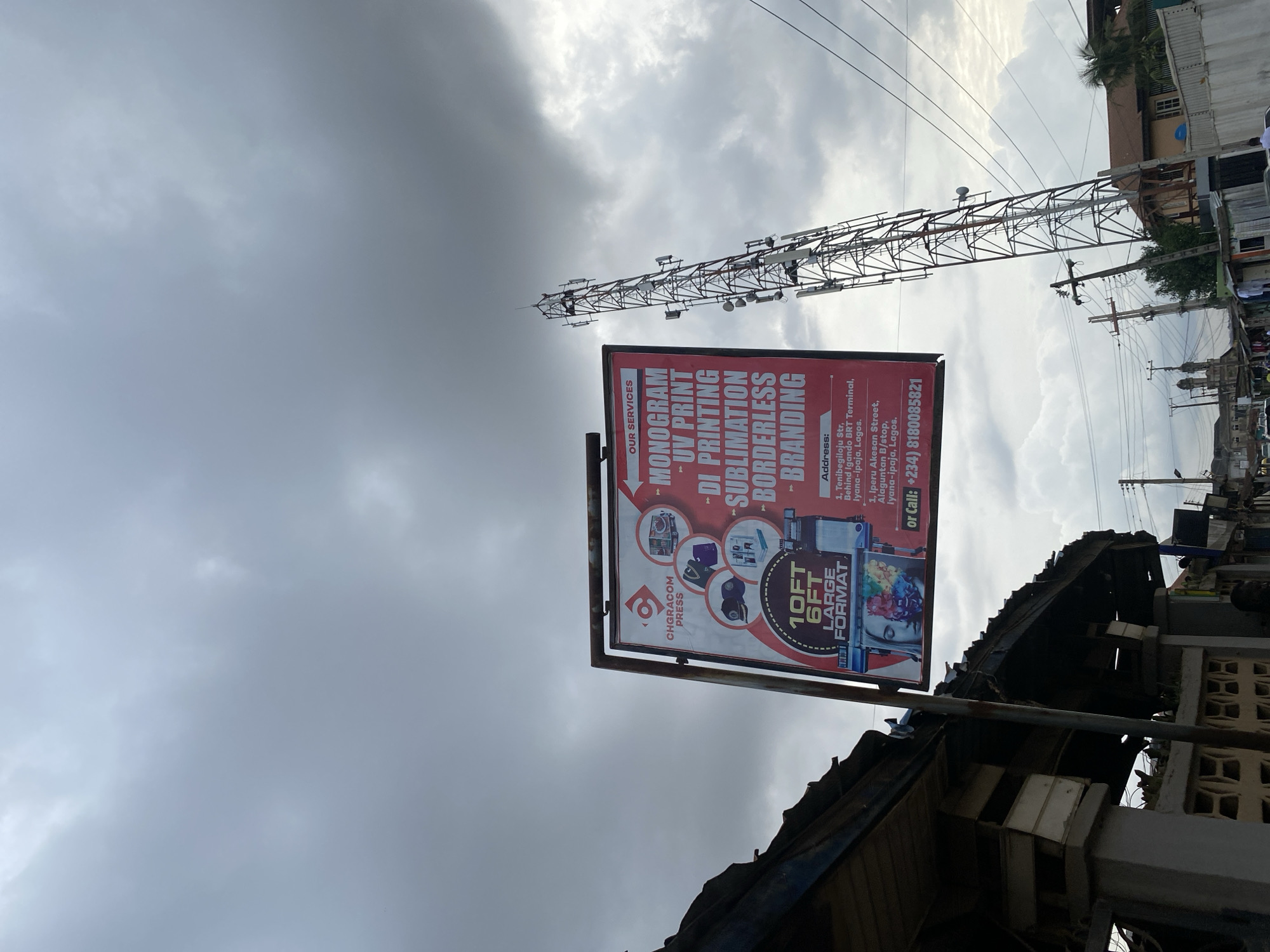 The image shows an advertisement sign for "Chgracom Press" located in Lagos, Nigeria. The sign is red and advertises monogram, UV print, DI printing, sublimation, borderless, and branding services. The sign lists two addresses: "1, Tenibegiloju Str, Behind Igando BRT Terminal, Olyana-ipaja, Lagos" and "1, Iperu Akesan Street, Alaguntan B/stop, Iyana-ipaja, Lagos." A telephone tower is visible in the background. The sign is attached to a structure with a dilapidated roof. The sky is cloudy.