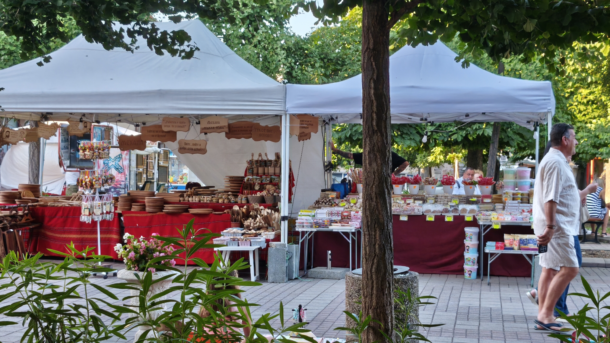 The image depicts an outdoor market scene in Pomorie, Bulgaria, under a sunny sky. Two white canopy tents are set up, providing shade for stalls displaying various goods. The stall on the left is laden with wooden kitchenware, including plates, bowls, and utensils, arranged on red fabric. Decorative items and jars of preserves are also visible. The stall on the right features an assortment of candies, sweets, and packaged food items, presented in colorful containers and on shelves. In the foreground, lush green plants are in focus, with a tree trunk partially obscuring the view. Several people are present in the market. A man in a light-colored shirt and shorts is walking past the stall on the right, holding a phone. Another person, partially visible in the background, appears to be a vendor. The market is situated on a paved area, with trees and foliage contributing to the natural setting.