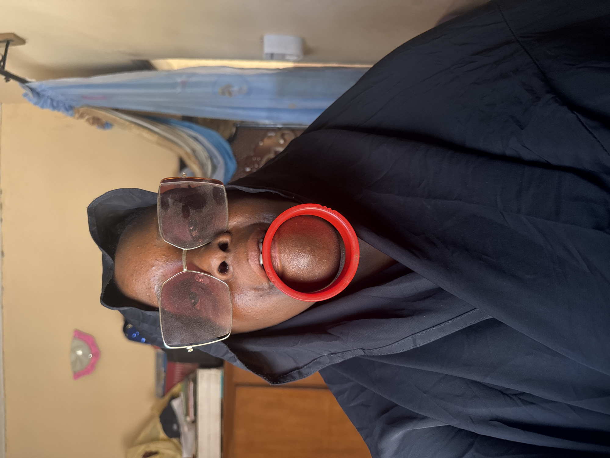 A woman wearing eyeglasses, a black garment, and a large red lip plate is depicted in this indoor, medium shot. The location is potentially Madangala, Nigeria. The woman is oriented vertically, with her head tilted slightly to the left. The eyeglasses have a thin metal frame and rectangular lenses. The lip plate is red and circular, stretching her lower lip significantly. The black garment covers her head and drapes over her shoulders.
The background includes a wall painted in a light brown tone. In the upper portion of the image, a mosquito net with blue trim is visible. A few books are visible behind the woman.