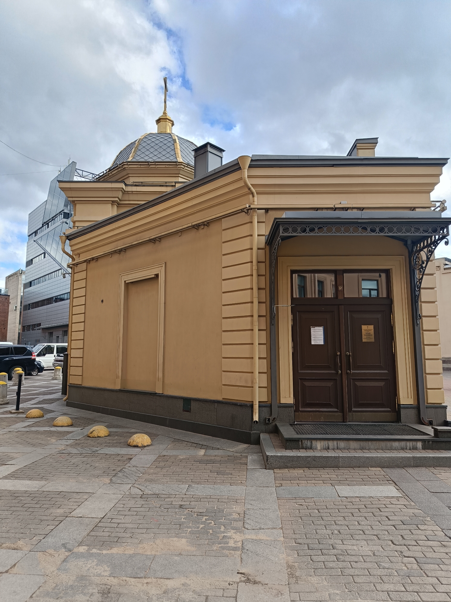 The image depicts a small, one-story building in what appears to be an urban setting. The building is painted in a yellow-beige color with dark brown accents. It features a double door entrance with a decorative metal canopy. A dome is visible on top of the building, with a cross on its peak. The building has a closed-off entrance on its side. The street is paved with grey bricks. There are yellow spherical bollards on the side of the building. In the background, a modern, multi-story building with a glass facade is visible. The sky is cloudy and overcast. The location is Unknown, Unknown.