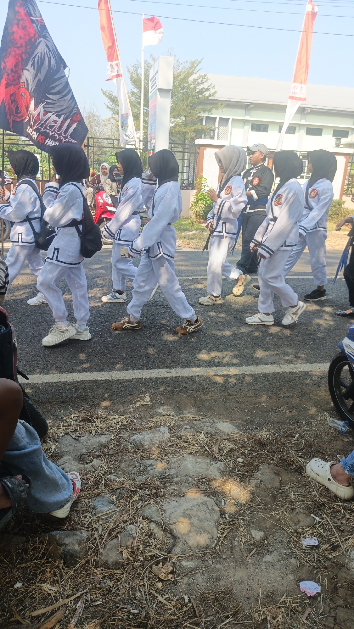 In Kandangmas, Indonesia, a group of individuals, predominantly young women wearing white uniforms and headscarves, are participating in a parade. They are walking on a paved road, with some wearing black sashes or bags. A large flag with red and black graphics, possibly a logo or emblem, is held aloft by a person at the front left of the group. Behind them, other flags, including the Indonesian flag (red and white), are visible. In the background, a building with a light-colored facade and windows can be seen, along with trees and a clear sky. The ground in the foreground is covered with dry grass, dirt, and rocks. Other people, some seated and some standing, are visible on the sides of the road, observing the parade. A red motorcycle is parked to the left of the parading group.