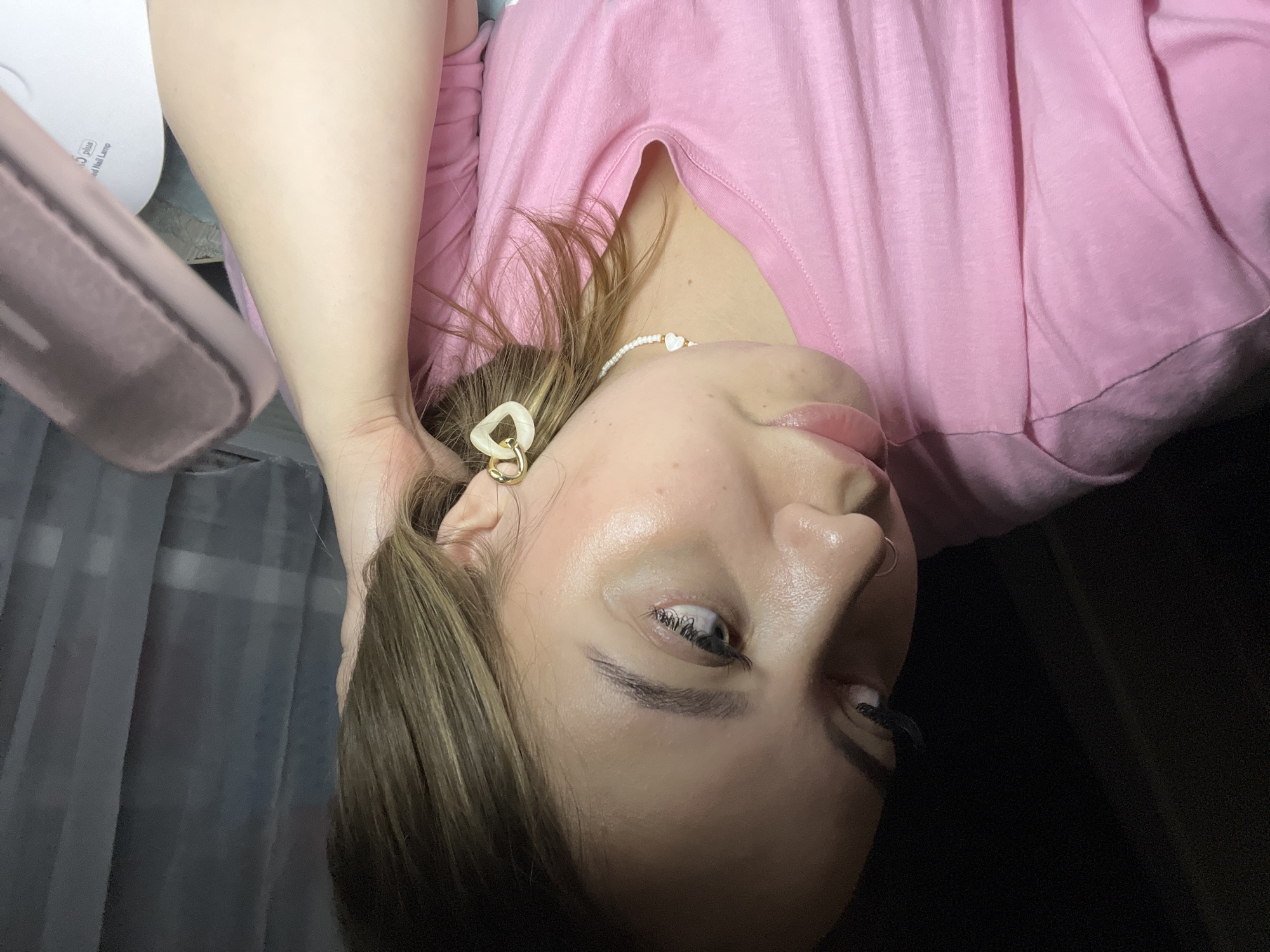 A woman with light brown hair and fair skin is wearing a pink t-shirt. She has a gold and white earring in her left ear. She is also wearing a white beaded necklace with a heart pendant. Her left hand is holding her head. The image was not taken in Novopokrovskaya, Russia.