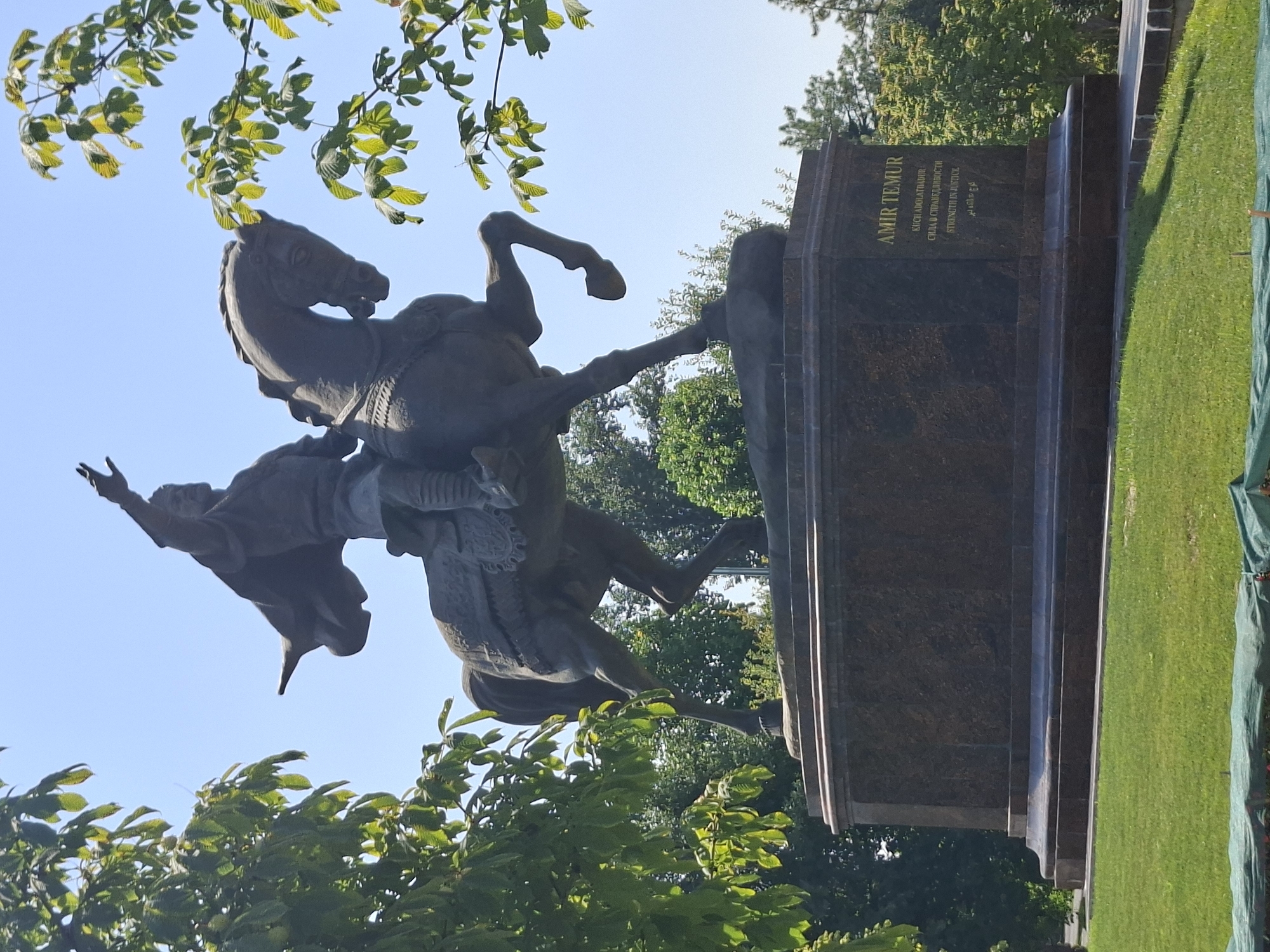 This is a low-angle, full-shot photograph of a bronze equestrian statue of Amir Temur, located in Amir Temur Square, Tashkent, Uzbekistan. The statue depicts Amir Temur on horseback, with his right arm raised in a commanding gesture. The statue is set on a large, multi-tiered granite pedestal, which has an inscription in golden lettering on its right side that reads "AMIR TEMUR". The inscription is in Cyrillic and Latin scripts.
The statue is positioned in a park-like setting with lush green grass in the foreground and trees surrounding the pedestal. The sky is a clear blue, indicating a sunny day. The lighting suggests that the photograph was taken during the daytime. The statue is the central focus of the image, and the surrounding environment provides context for its placement. The image captures a sense of historical significance and grandeur associated with the figure of Amir Temur.