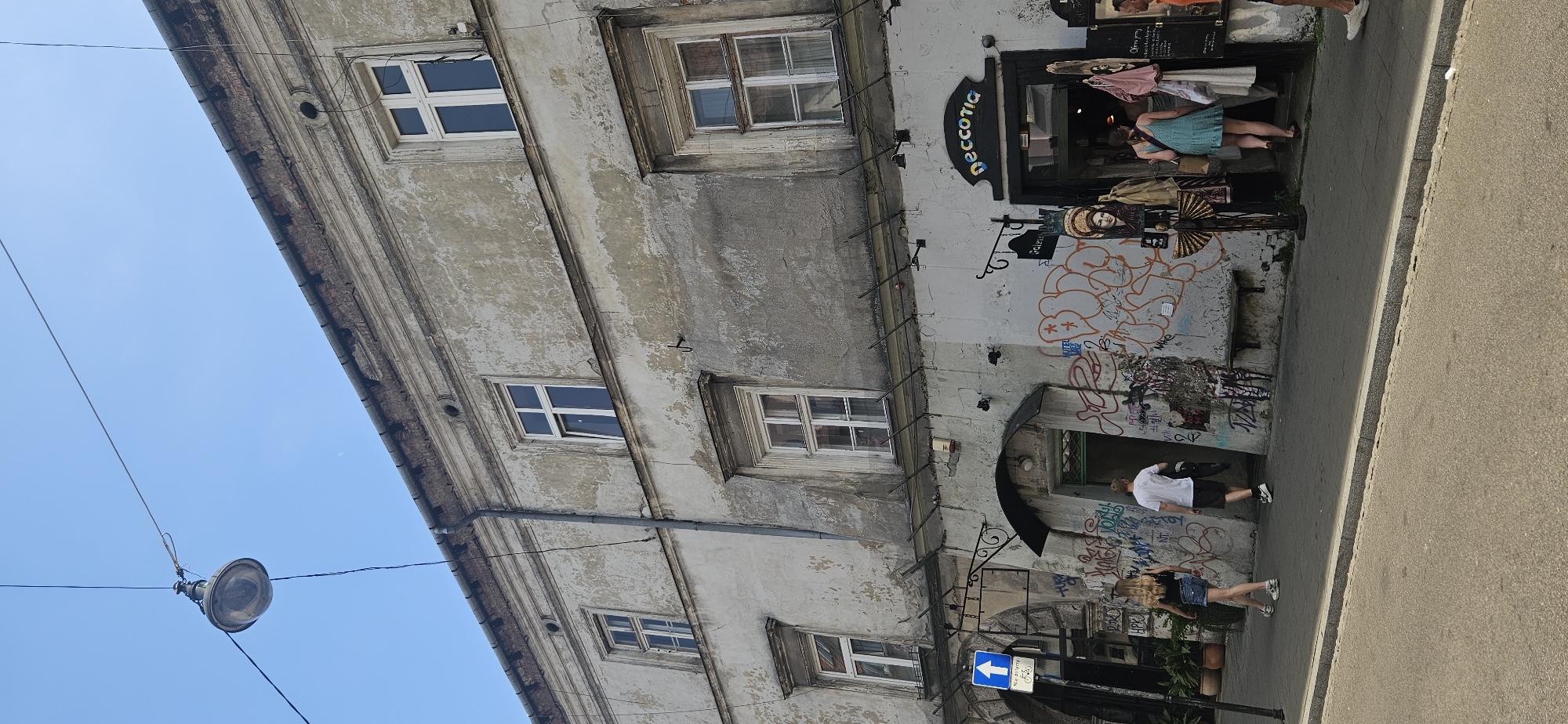 The image depicts a street scene in Krakow, Poland. A weathered, multi-story building with multiple windows dominates the upper portion of the frame. The facade is peeling and shows signs of age. In front of the building, a street level business is visible with a sign that reads "NECCOTIA" in colorful lettering. The entrance to the business is open, and several people are entering and exiting. The walls of the building at street level are covered in graffiti. A sidewalk runs along the front of the building, and a street is in the foreground. There are a few people walking on the sidewalk, including a woman in a black top and denim shorts, and a man in a white t-shirt and shorts. A blue traffic sign with a white arrow is visible on the left side of the image. The sky is clear and blue.