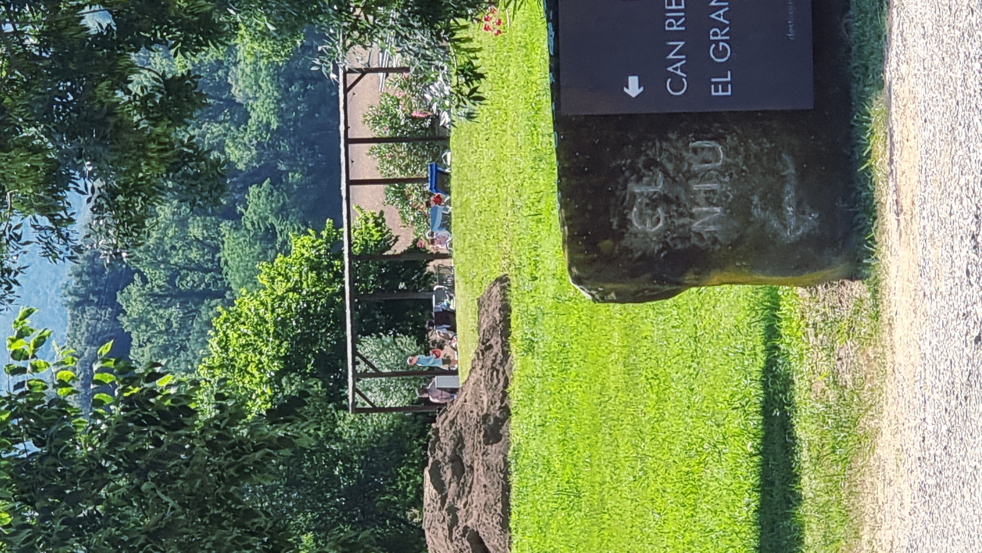 In Montagut i Oix, Spain, an outdoor scene unfolds with a focus on a grassy area and a shaded pavilion. A large mound of dark soil is prominent on the left side of the frame. Several individuals, including adults and children, are gathered under a wooden structure with a brown roof, some sitting on chairs and others appearing to be interacting. The ground is covered in lush green grass, with some patches of dry earth. In the foreground, a rough-hewn stone stands to the right, bearing carved inscriptions. A dark sign is affixed to this stone, featuring a white arrow pointing left and text in white lettering, partially visible as "CAN RIE" and "EL GRAN". The background is a dense forest with trees of varying shades of green, leading to a distant, hazy mountain range. Sunlight dapples through the leaves of trees in the foreground, casting shadows on the grass.