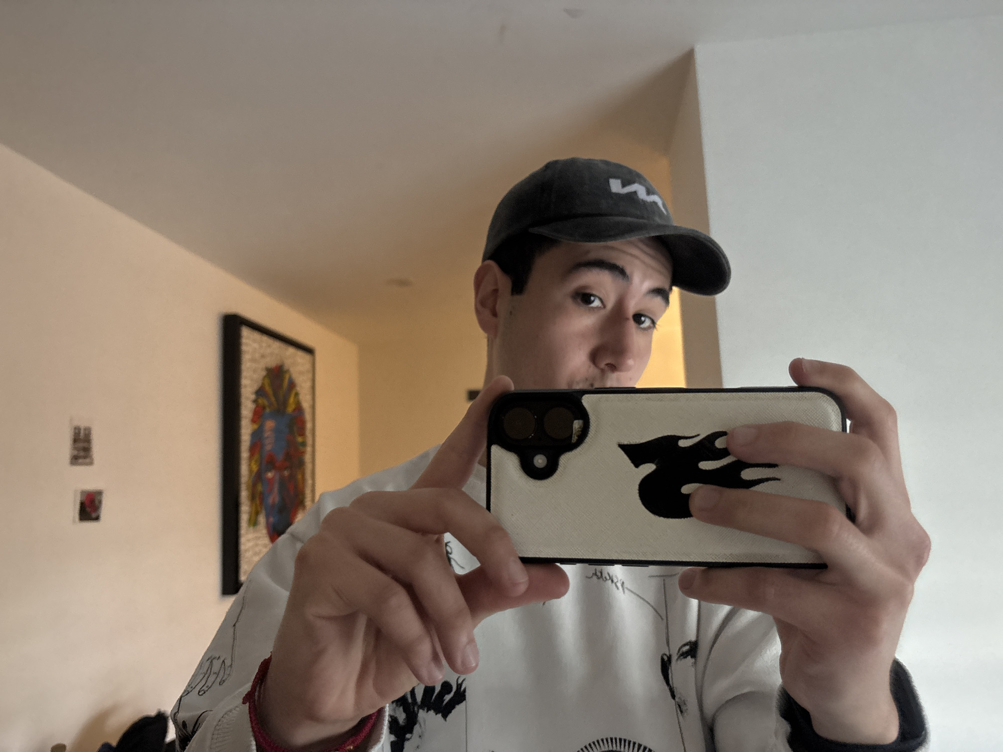 A young man in San Bernardino Tlaxcalancingo, Mexico, is depicted taking a selfie indoors, likely in a mirror. He faces forward, looking directly into the camera with a neutral expression. He wears a dark gray baseball cap featuring a light-colored, wavy logo, possibly a stylized "W" or "M", on its front. His attire includes a light-colored (likely white) sweatshirt adorned with black graphic designs that appear to be abstract figures or sketches. A red braided or beaded bracelet is visible on his left wrist.
He holds a smartphone horizontally with both hands. The phone is fitted with a light cream or off-white case that prominently displays a black flame-like or stylized "S" design on its back. The phone's dual camera lenses are visible on the top left of the case.
The background consists of light-colored walls and a ceiling. On the left, a large framed artwork is visible, portraying a colorful, mosaic-like figure in vibrant reds, blues, and yellows. Below this main piece, two smaller, darker framed items hang on the wall. The bright lighting suggests a well-lit indoor environment, possibly during the daytime.