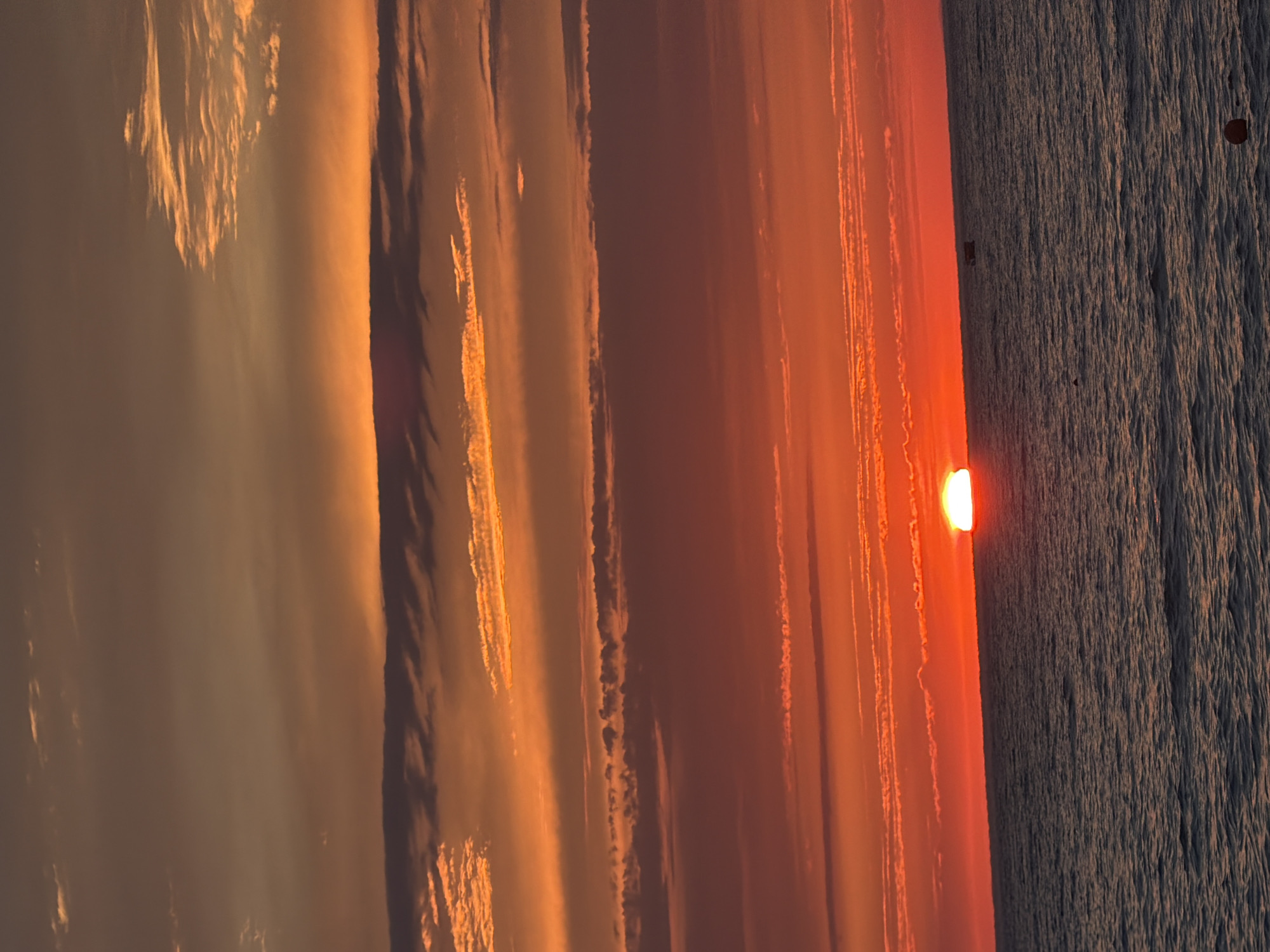 The image depicts a serene sunset over a body of water, likely the sea, given the context of Sochi, Russia. The sun, a vibrant orange orb, is partially submerged at the horizon, casting a warm, fiery glow across the sky and water. Wispy clouds, illuminated by the setting sun, stretch horizontally, creating streaks of light and shadow that add depth and texture to the scene.
The water's surface is textured with ripples, suggesting a gentle breeze or subtle currents. A thin, dark line along the horizon separates the sky from the water, emphasizing the vastness of both elements. Small, dark objects are visible on the water, possibly boats or buoys, adding a touch of scale to the immense landscape.
The overall mood is one of tranquility and natural beauty, characteristic of a sunset in a coastal location. The time of day is clearly evening, as indicated by the low position of the sun and the rich, warm colors painting the sky. The lighting is soft and diffused, creating a peaceful and contemplative atmosphere. No text is visible in the image.