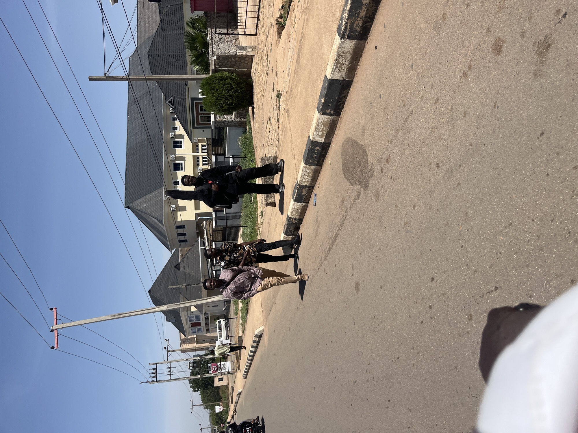 The image depicts a sunny street scene in Gura Top, Nigeria. Three men are walking along the street, which has a paved surface and a curb with black and white markings. The man closest to the camera wears a checkered shirt and khaki pants, while the other two are dressed in darker clothing, including one in all black. Buildings are visible in the background, along with utility poles and wires. A motorcycle is parked nearby.