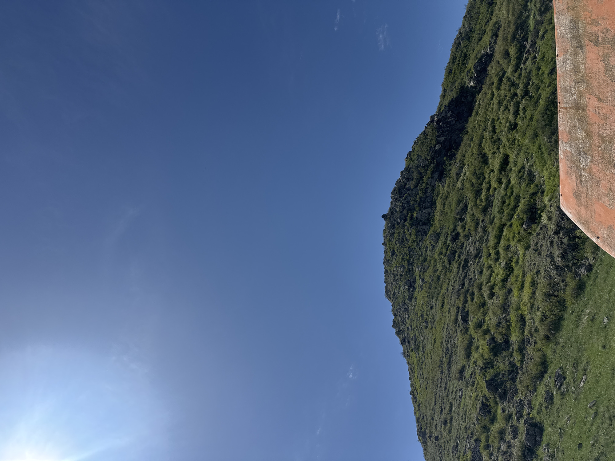 The image shows a landscape with a clear blue sky covering most of the left side. A mountain covered in green vegetation rises on the right side. A rust-colored, textured structure is visible at the far right edge of the image, partially obscuring the mountain. The location is Bagravan, Armenia. No people are visible. The time of day appears to be sunny with bright daylight.