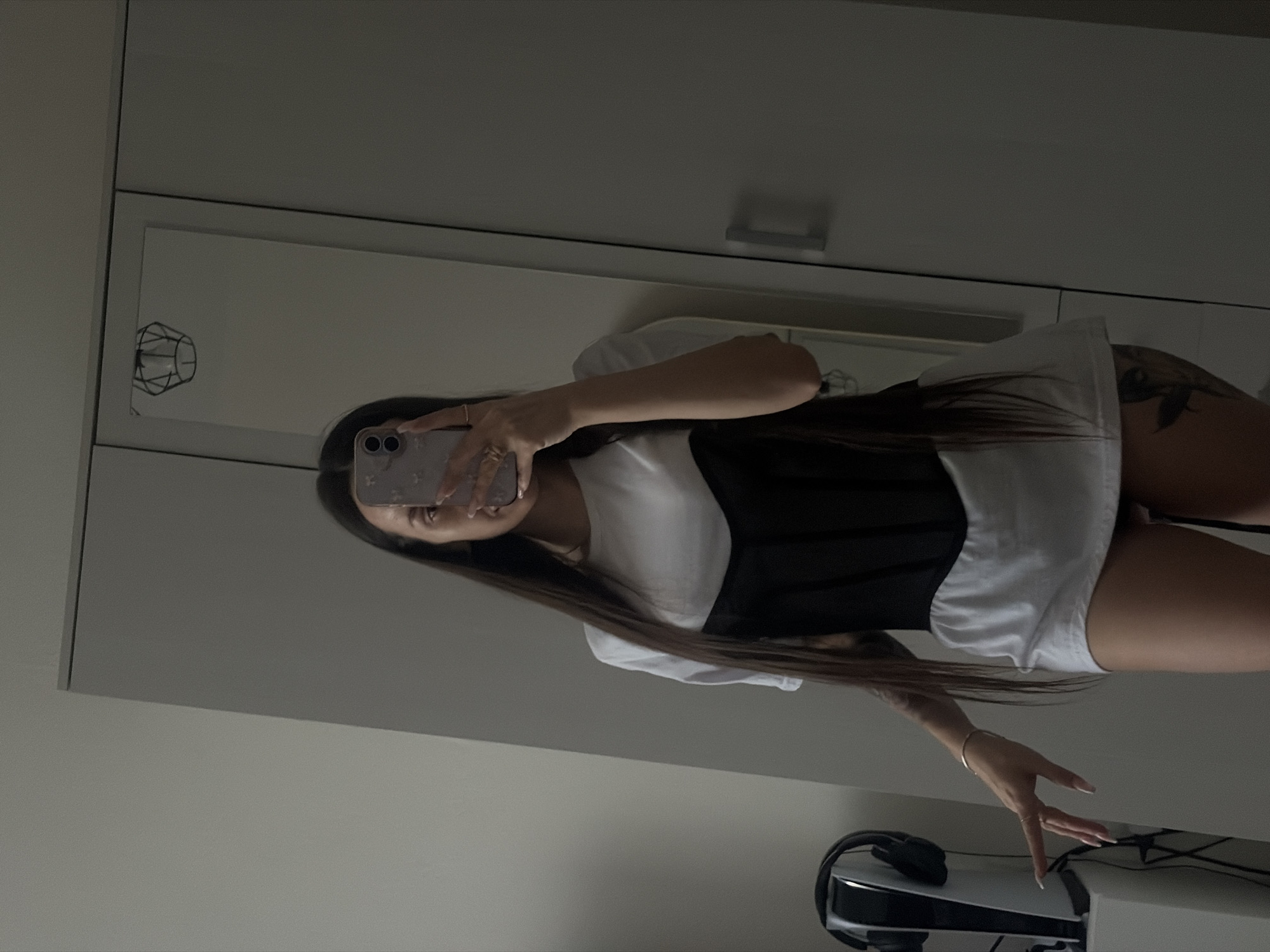 The image shows a person taking a selfie in front of a mirror. The person has long, dark hair and is wearing a white t-shirt with a black corset. The person is holding a phone in their left hand. The background shows a closet with a mirrored door, a decorative light fixture, and a PlayStation 5. The location is not identifiable as Saint Petersburg, Russia.