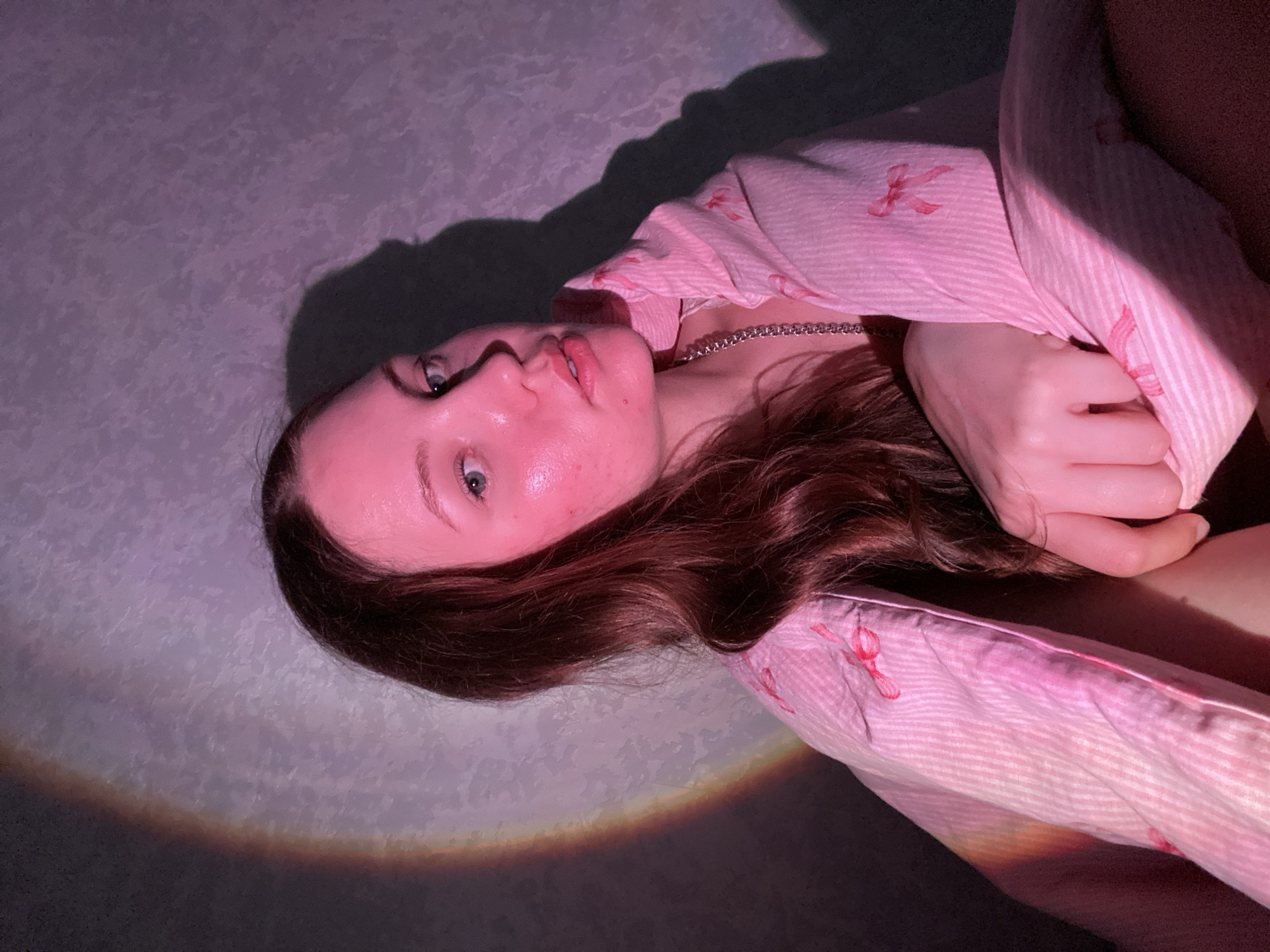 A young woman with long, wavy brown hair is looking towards the camera, partially wrapped in a pink striped blanket adorned with small pink bows. She is wearing a silver chain necklace. The lighting in the scene is dramatic, with a pinkish hue cast over her skin and a distinct shadow on the wall behind her. A rainbow-like light effect is visible on the left side of the frame, casting colors onto the wall and the blanket. The background appears to be a textured wall. There is no indication of the specific location within Izhevsk, Russia.