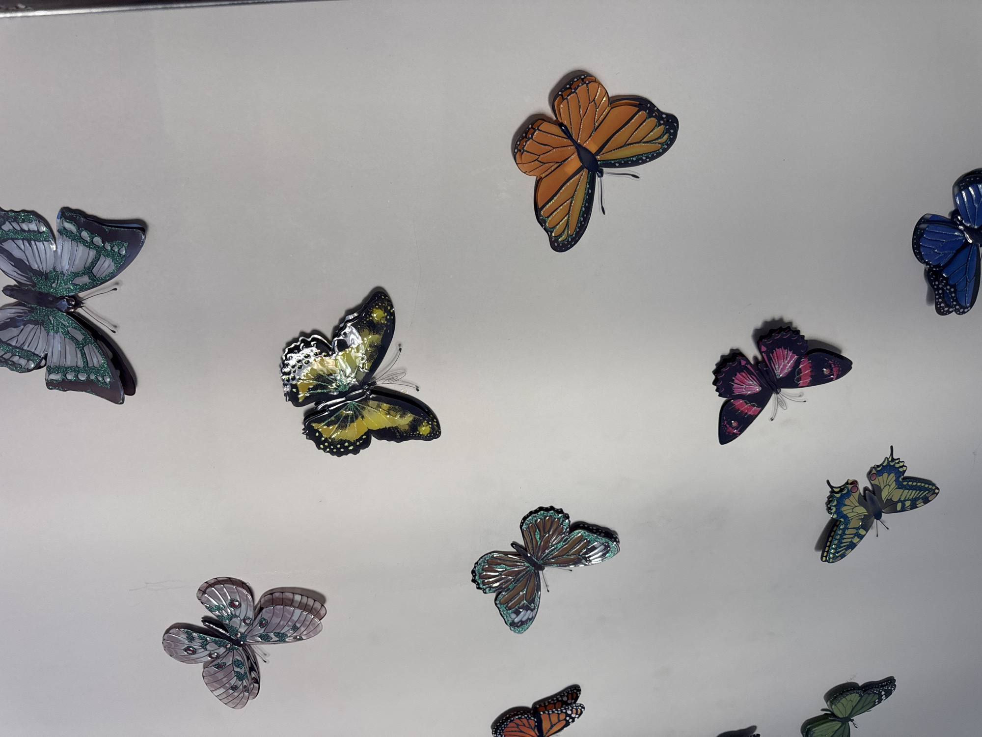 The image displays a collection of decorative butterflies attached to a light-colored surface, possibly a wall or ceiling. The butterflies are varied in color and pattern, with some featuring iridescent or metallic finishes. They appear to be made of paper or a similar craft material, with some having slightly raised wings to give a three-dimensional effect. The arrangement is somewhat scattered, suggesting a decorative application rather than a natural habitat. There are at least ten visible butterflies of different sizes and colors including blue, orange, green, purple, and yellow. The lighting suggests an indoor environment, and the surface is plain and uniform. No people, text, or specific location cues beyond the general context of decorative items are visible.