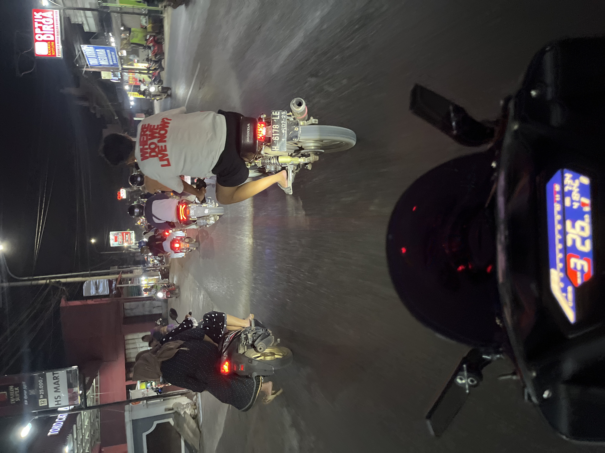 The image captures a nighttime scene on a street in Kalinyamatan, Indonesia, focusing on a group of people riding motorcycles. In the foreground, the dashboard of a motorcycle is visible with a digital display showing "3 26." Behind this, the rider of another motorcycle is seen from behind, wearing a white t-shirt with red text that reads "WHERE DO WE LIVE NOW?". This rider is on a Honda motorcycle with the license plate "H-6178-LE" and a sticker showing "20-07-25". Several other motorcycles and riders are also on the road, some with passengers. The environment appears to be a commercial area with buildings and streetlights illuminating the scene. Signs for businesses like "HS MART," "DIARLIM SUPER," and "OPTİK DİRGA" are visible on the buildings. The road surface shows reflections from the lights, suggesting it might be wet.