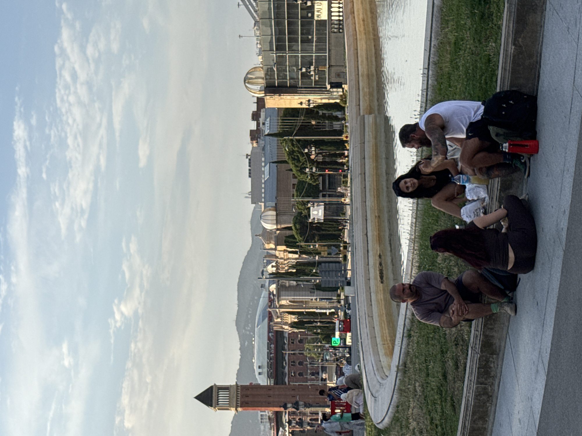 In Barcelona, Spain, on a bright, overcast day, four individuals are gathered near a fountain, engaging in conversation. The scene is set against a backdrop of urban architecture, including a prominent clock tower, various buildings, and distant hills.
In the foreground, two men and two women are seated on the grass or the paved walkway. The man on the left is wearing a brown t-shirt, dark shorts, and green socks. He is sitting with his legs crossed, his hands clasped in front of him. The woman next to him is seated, facing away from the camera, wearing a dark top and dark bottoms.
To the right, a woman with dark hair is seated, wearing a black tank top and what appear to be dark shorts. She is looking towards the man beside her, who is wearing a white tank top and dark shorts. This man has tattoos on his arms and is gesturing with his right hand. A red thermos and a backpack are near their feet.
The fountain in the mid-ground features a curved edge with a shallow body of water. Beyond the fountain, the cityscape unfolds. A tall, brick clock tower with a pyramidal top stands to the left, with lampposts in front of it. Behind the tower are several buildings, some with arched windows and domes, and a large, modern building with a glass facade. Trees are scattered throughout the urban landscape. In the distance, mountains are visible under a cloudy sky. A red bus is seen on the street in the background, alongside other vehicles and people walking.