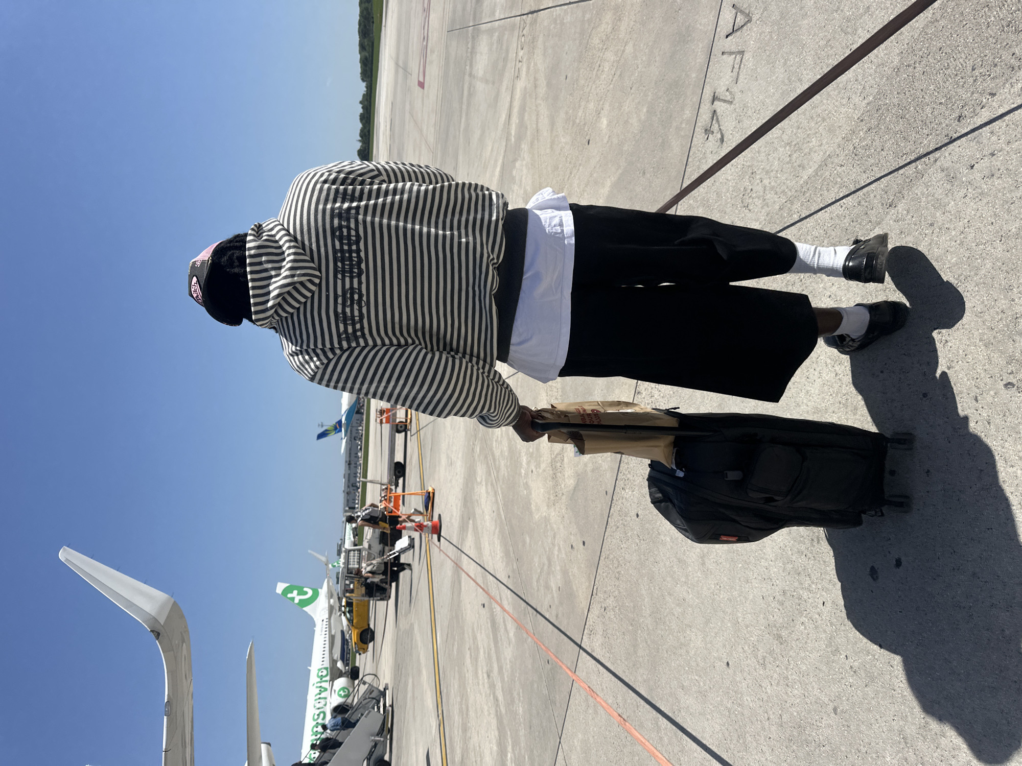 The image shows an individual standing on an airport tarmac. The person is wearing a black and white striped hooded sweatshirt, a white undershirt, black pants, white socks, and black shoes. They are pulling a black wheeled suitcase with a brown item resting on top. The tarmac has various markings and the letters "AF14" are visible. In the background, there are airplanes, including one with the Transavia logo. The location is likely an airport within Paray-Vieille-Poste, France.