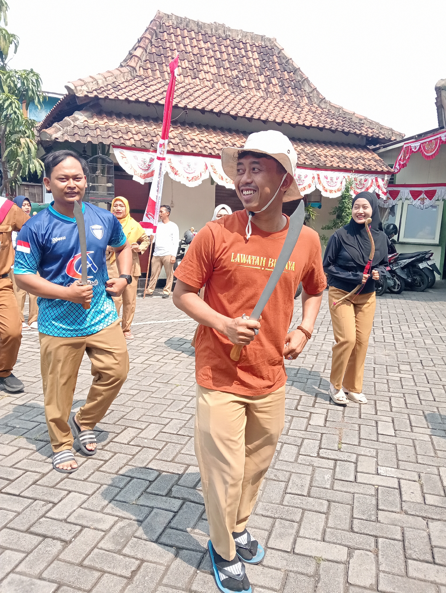 The image depicts a sunny outdoor scene in the city of Demak, Indonesia, likely a public space given the paved brick ground and traditional tiled-roof buildings in the background. In the foreground, a man in an orange t-shirt with "LAWATAN BI...A" printed on it, wearing a white bucket hat and tan pants, is joyfully holding a machete. To his left, another man in a blue and light blue patterned t-shirt and tan pants also holds a machete. Behind them, a woman in a yellow headscarf and tan pants stands, and further back, a person in a white shirt and tan pants holds a red and white flag. To the right of the central figure, a woman wearing a black hijab and black top over tan pants smiles while holding a bow and arrow. Several motorcycles are parked behind a building to the far right. The overall atmosphere suggests a festive event or performance.