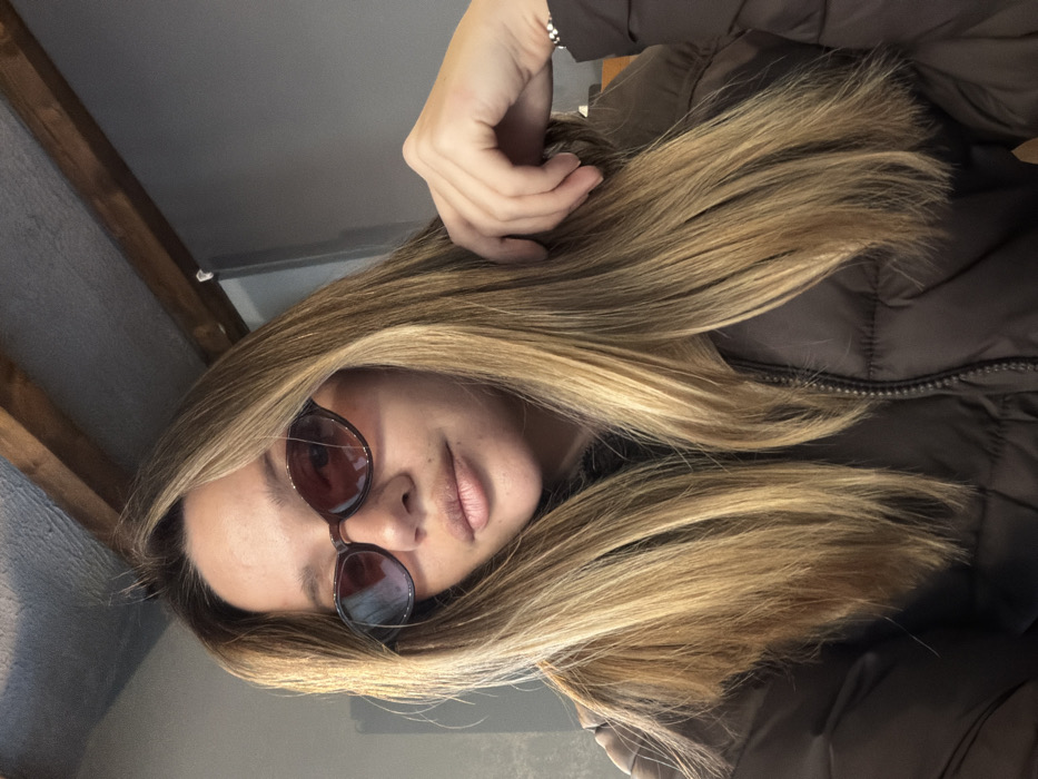 A female subject is depicted in a frontal, medium-close-up indoor shot. She has mid-length, straight light brown hair with blonde highlights, flowing over her shoulders. She wears dark brown, oval-shaped sunglasses with reddish-amber translucent lenses, which show reflections of a blue light source. Her lips are slightly parted in a neutral expression. She is dressed in a dark brown puffer jacket with a visible front zipper. Her left hand, adorned with a silver-colored bracelet on the wrist, is positioned near her hair on the right side of the frame. The background features a light grey ceiling area, with two dark wooden beams running horizontally across the upper right section, and a plain grey wall to the right. The setting is indoors, city of Unknown, Unknown.