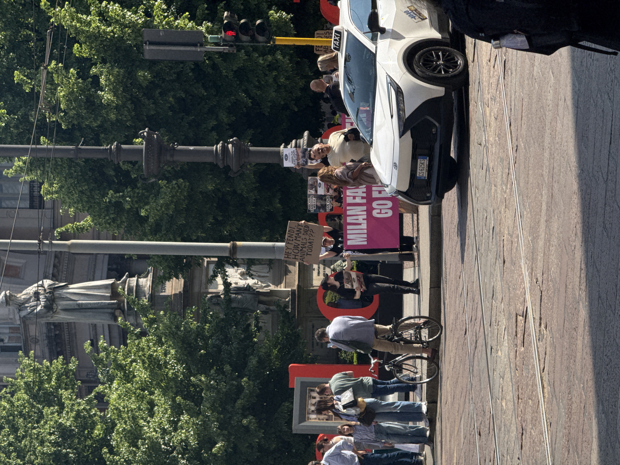 In Milan, Italy, a street scene reveals a gathering of individuals holding signs, likely engaged in a protest or demonstration. The banners bear messages such as "MILAN FASHION WEEK" and "BAN FUR NOW!". One sign reads, "HEY FENDI HOW MANY ANIMALS DIED TODAY??". A white taxi cab with a “TAXI” sign is positioned on the side of the road. People are standing on the street, some with bicycles. In the background, a statue is visible behind some trees. A traffic light with a red light is also visible. The street is paved with stone.