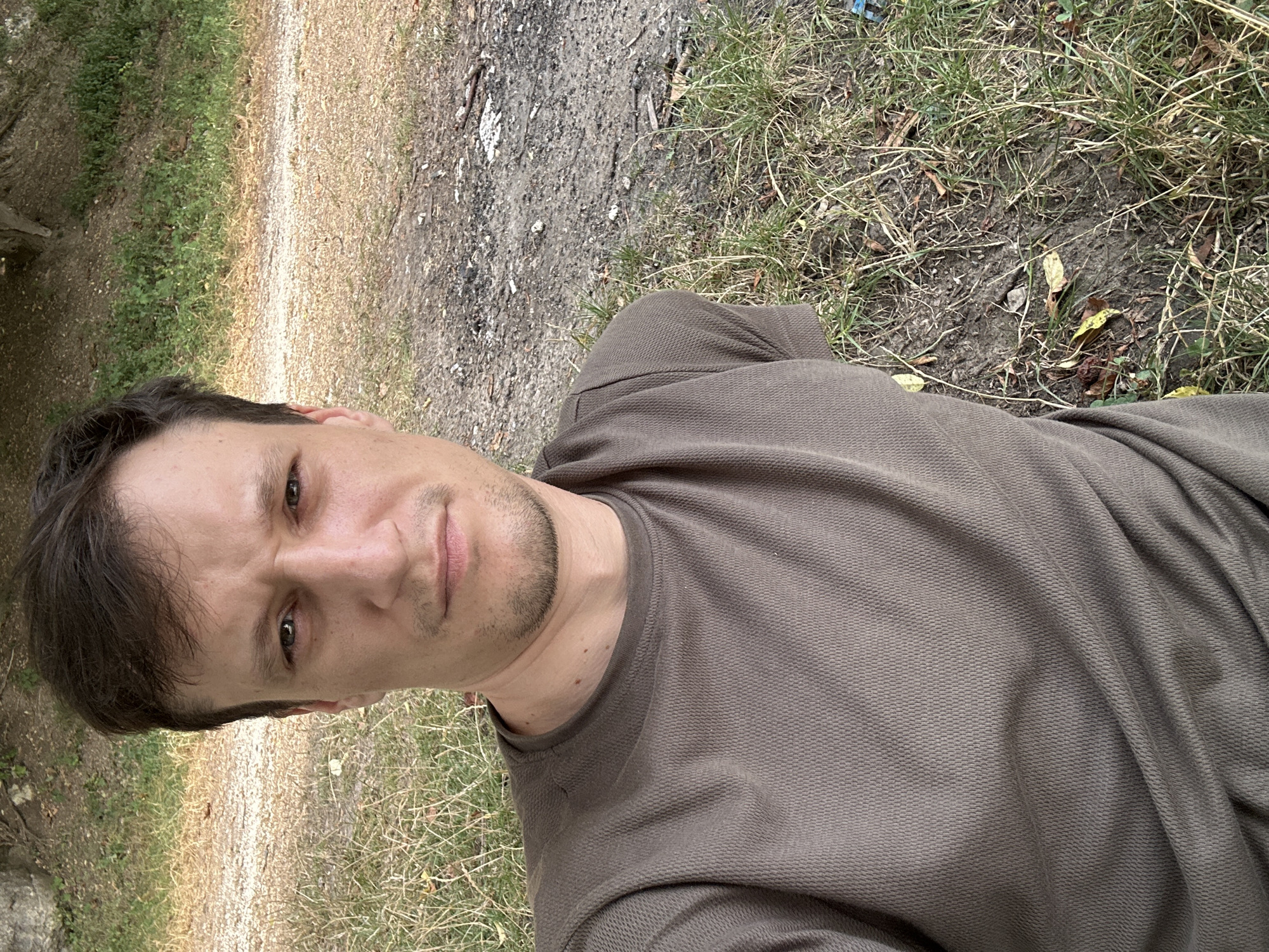 This is a selfie taken by a man outdoors, likely in a park or wooded area in Makiv, Ukraine. The man is wearing a short-sleeved, crew-neck t-shirt of a dark olive color. He has short, dark brown hair and a light stubble on his face. He is looking directly at the camera with a neutral expression. The background consists of dry grass, dirt, and some scattered leaves, with a hint of trees and foliage in the upper portion of the image, suggesting a natural setting. The lighting appears to be natural daylight.