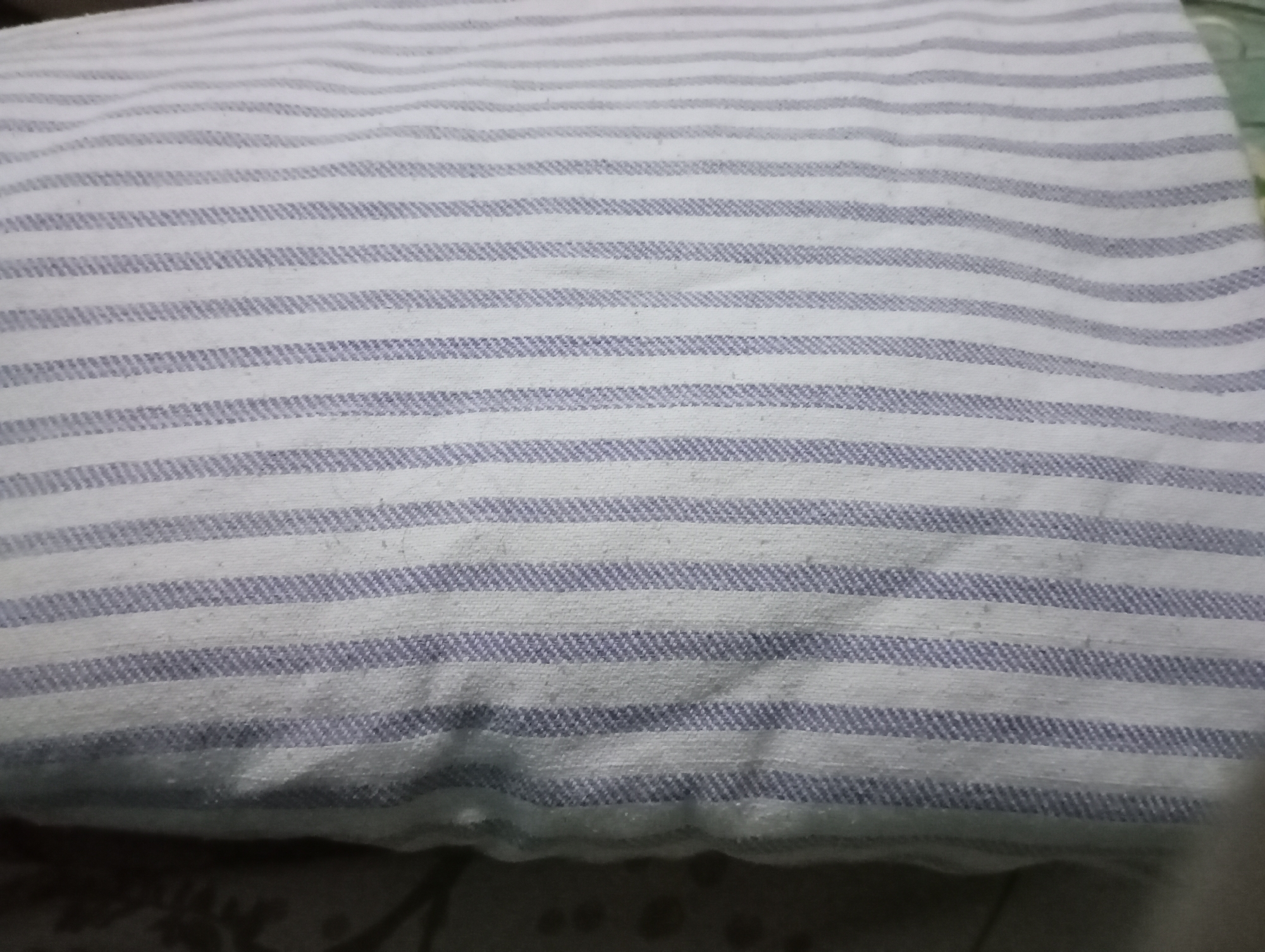 The image displays a close-up view of a white fabric with thin, horizontal lavender stripes. The fabric appears to be folded, creating soft curves and shadows across its surface. There are some small, light-colored specks visible on the fabric, suggesting it might be slightly worn or have lint. The background is dark and out of focus, with some faint patterns visible at the bottom, possibly indicating a surface or another object beneath the fabric. The scene is devoid of any human presence or discernible environmental context that would place it specifically in Kecamatan Haurgeulis, Indonesia.