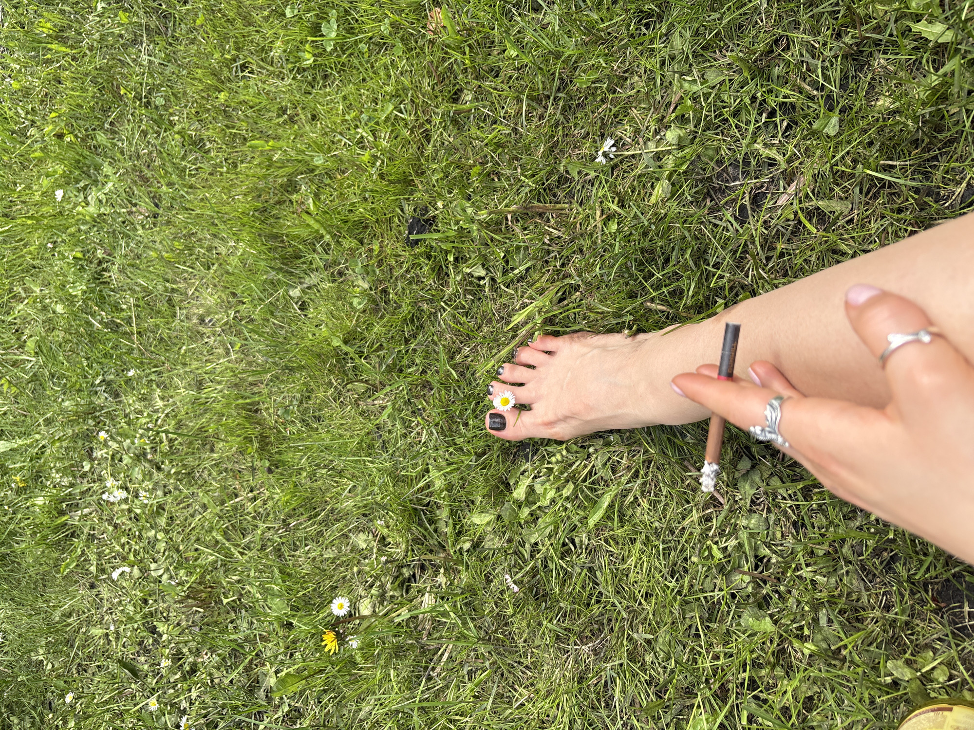 A partial view of a person's leg and hand are present in the image, set against a grassy background containing daisies. The person's toes are painted black, with a daisy placed between two of them. Their hand holds a rolled item, possibly a cigarette or herbal roll, adorned with a decorative tip. The person is wearing rings. The scene appears to be outdoors, in an area with green grass and wildflowers. The location is given as Пярэжырскі сельскі Савет, Belarus.