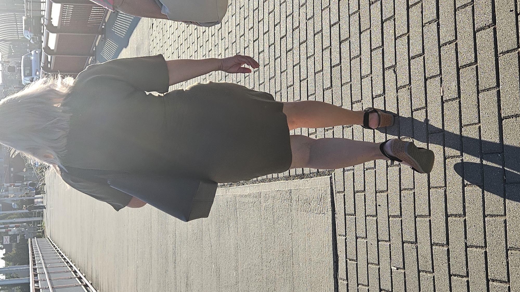 The image shows a woman walking away from the camera on a sidewalk made of brick pavers. She has blond hair and is wearing a dark olive green dress and wedge sandals. She is carrying a black shoulder bag. The sidewalk is bordered by a concrete path on the left and a metal railing on the right. In the background, there are some cars and buildings, suggesting an urban environment. The sun is shining, casting a long shadow from the woman on the pavement. There is no visible indication in the image that this location is in Prešov, Slovakia.