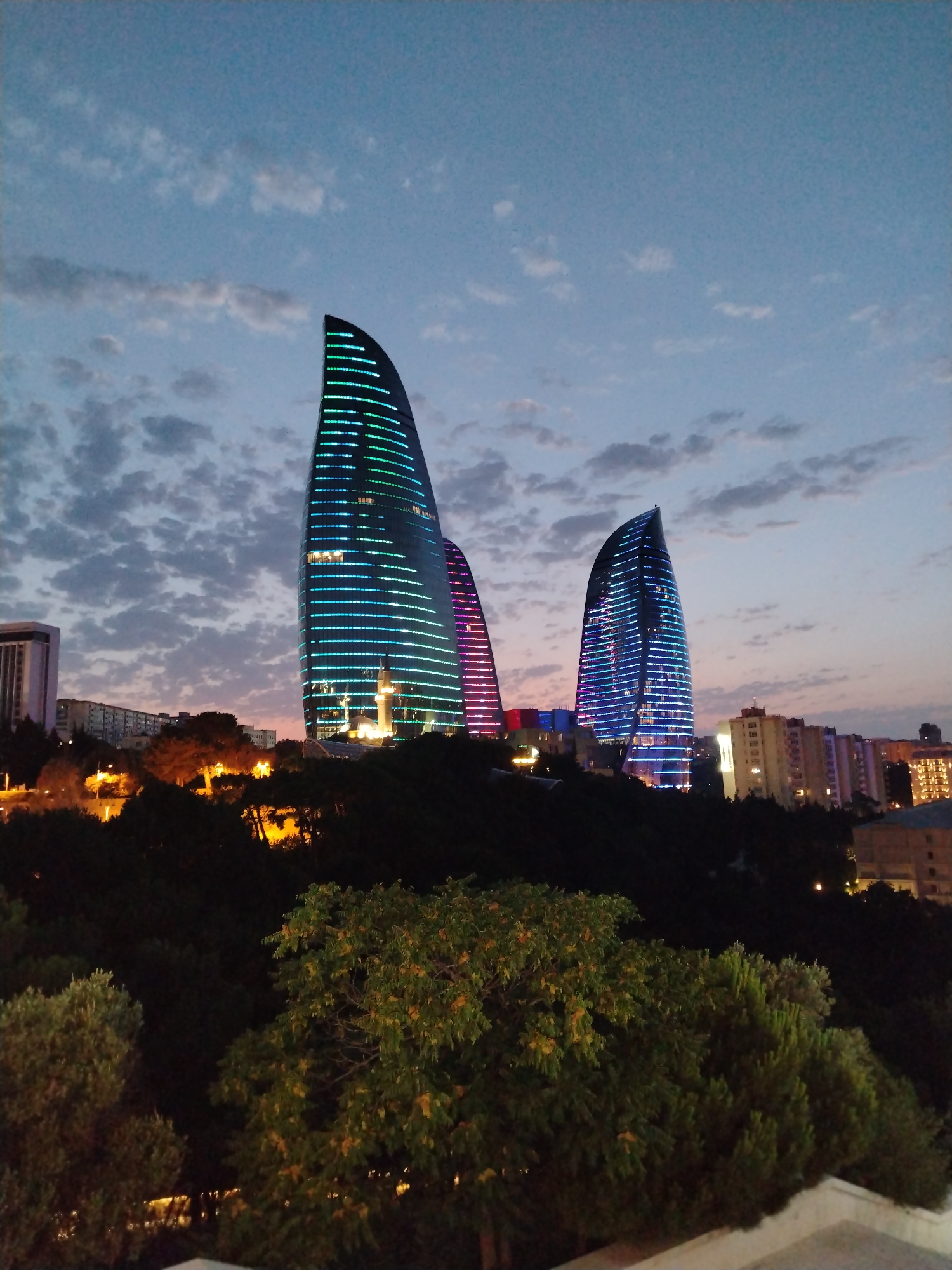 The image shows the Flame Towers in Sabail Raion, Azerbaijan, at dusk. The three flame-shaped skyscrapers are illuminated with colorful LED lights that mimic flames. The tallest tower is on the left and is lit with green and blue lights, while the middle tower is lit with pink and purple lights. The smallest tower is on the right and is lit with blue lights. In the foreground, there are trees and foliage, and in the background, there are other buildings and city lights. The sky is a deep blue with scattered clouds. A minaret of a mosque is visible between the Flame Towers.