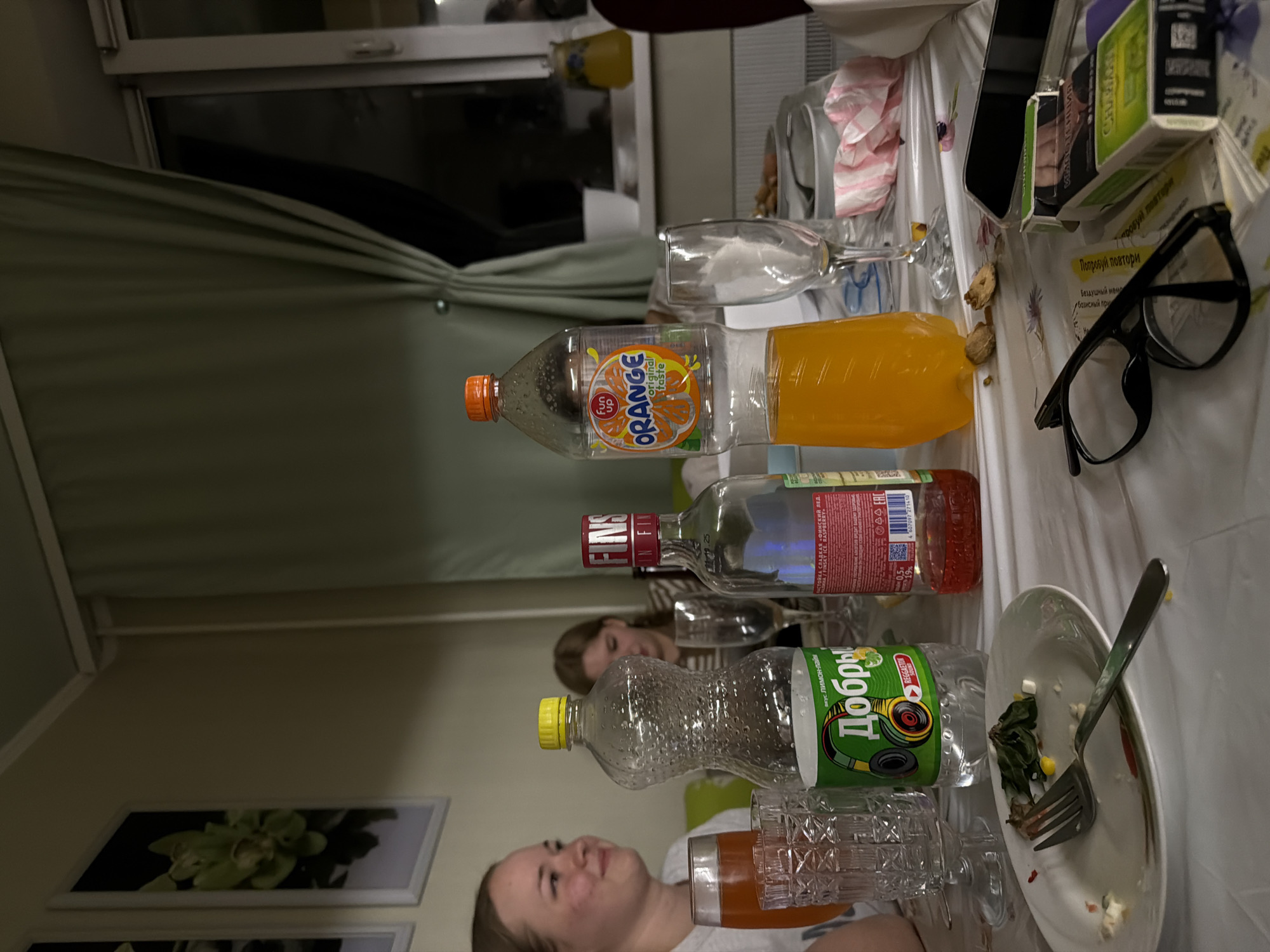 This indoor photograph, likely taken in Perm, Russia, captures a casual gathering around a table, suggesting an evening or nighttime setting due to the artificial lighting and dark view outside a window in the background. The image is oriented vertically but depicts a horizontal table.
Two young women are visible. In the foreground, a woman with light brown hair looks upwards with a faint smile, her features slightly soft-focused. Behind some bottles, another woman with darker hair is partially visible, observing something to the right.
The table is covered with a white tablecloth and is laden with various items. Prominently displayed are three plastic bottles: one containing orange juice labeled "ORANGE original taste," another with a clear liquid identified as "FINS ICE RASPBERRY" (a 0.5L, 19% alcohol sweet tincture), and a large "Добрый" (Dobry) bottle with "лимон-лайм" (lemon-lime) flavor. Several empty or partially filled glasses, including a wine glass and tumblers, are scattered around.
Remnants of a meal are present, including a white plate with leftover salad and a fork, and some scattered crumbs or food pieces on the table. Other personal items include a pair of black-framed eyeglasses, a black smartphone, and small packaged boxes. Text visible on these boxes includes "Попробуй повтори" (Try again) and partially "СИМПОТЕНЦИЯ" (Simpotentsiya) and "Бездушный мемл" (Soulless meme). A striped pink and white napkin is also on the table. In the background, part of a window with light-colored drapes or blinds is seen, and framed pictures of green orchids adorn the wall. The Cyrillic text on the labels confirms the Russian context.