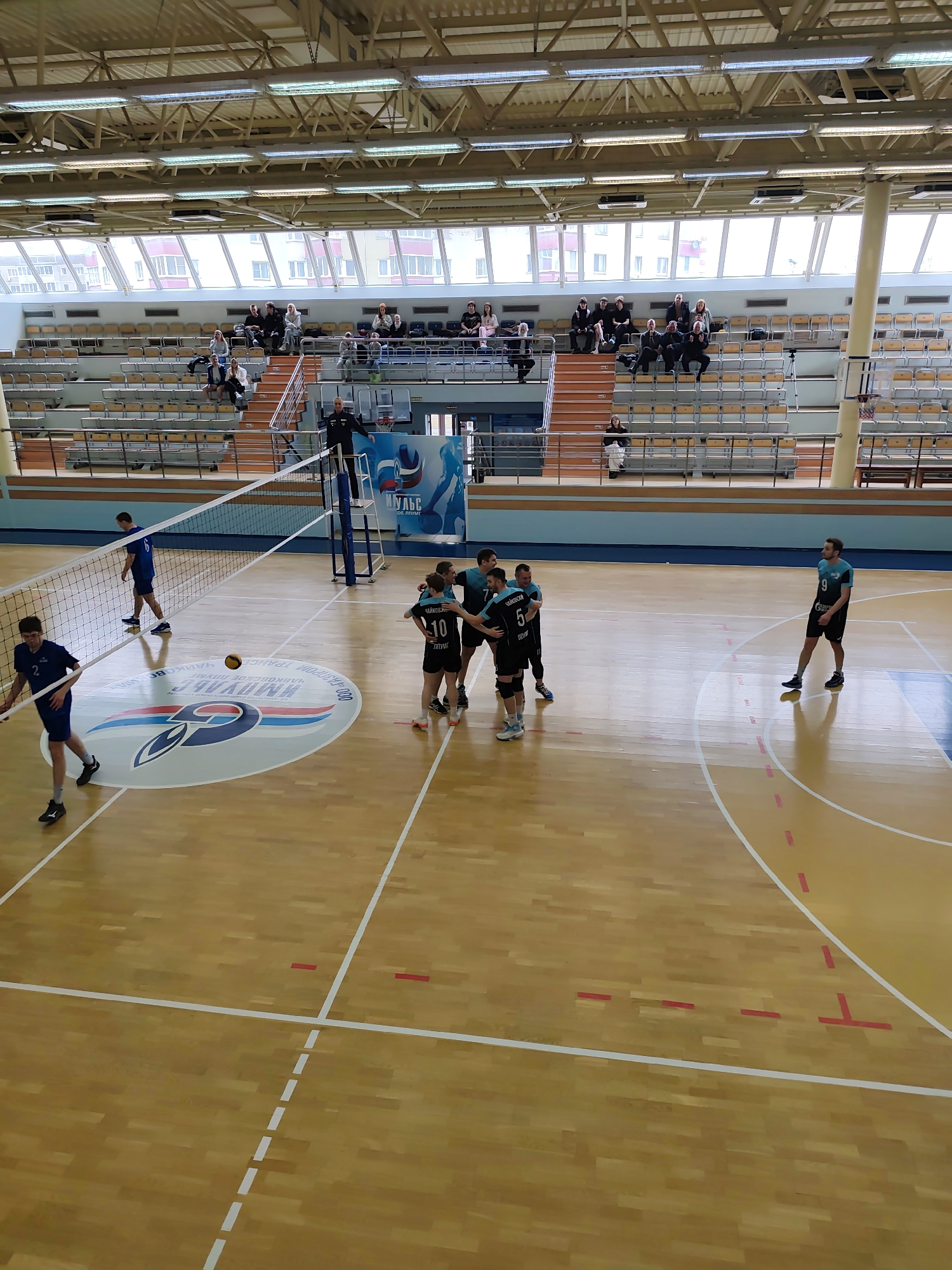 A volleyball game is underway in a gymnasium in Chaikovsky, Russia. The gymnasium features a light brown wooden floor with white lines marking the boundaries of the court. A volleyball net divides the court, with players from opposing teams positioned on either side. One team is celebrating, with four players clustered together in a group hug. They are wearing black jerseys with numbers on them. A player from the opposing team, wearing a blue jersey with the number 2, stands near the net, looking on. Another player from the opposing team, wearing a blue jersey with the number 6, is walking towards the net. A volleyball hangs in the air above the center of the court. In the background, rows of empty seats are visible in the stands. A person stands in a platform area between the court and the bleachers behind the net. People are seated in the upper rows of the bleachers. The gym is well-lit, with overhead fluorescent lights. The ceiling structure of the gym is visible, featuring exposed metal beams. A circular logo is printed on the floor of the court near the net.